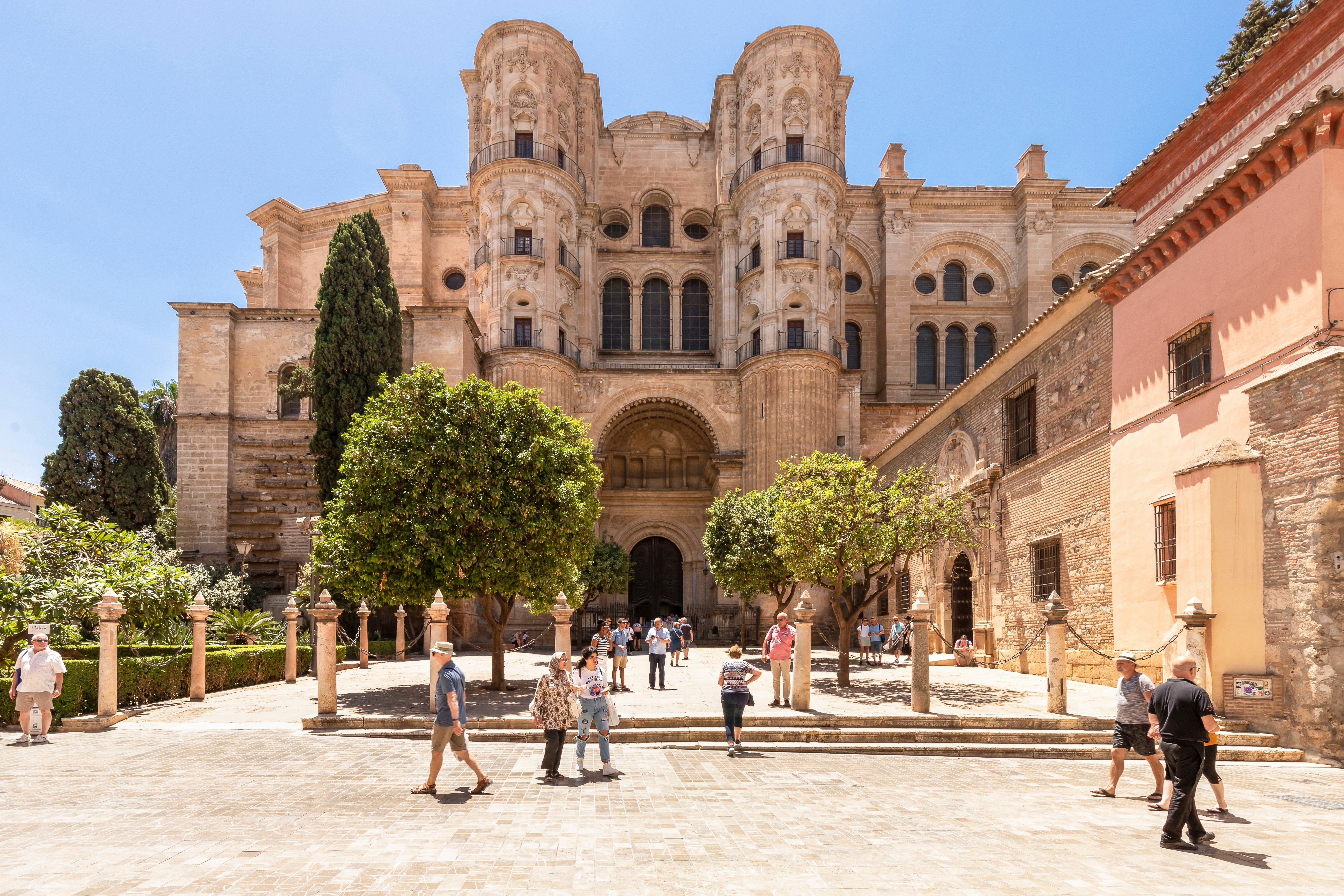 People walk around in a plaza with trees in front of a large, earth-toned building