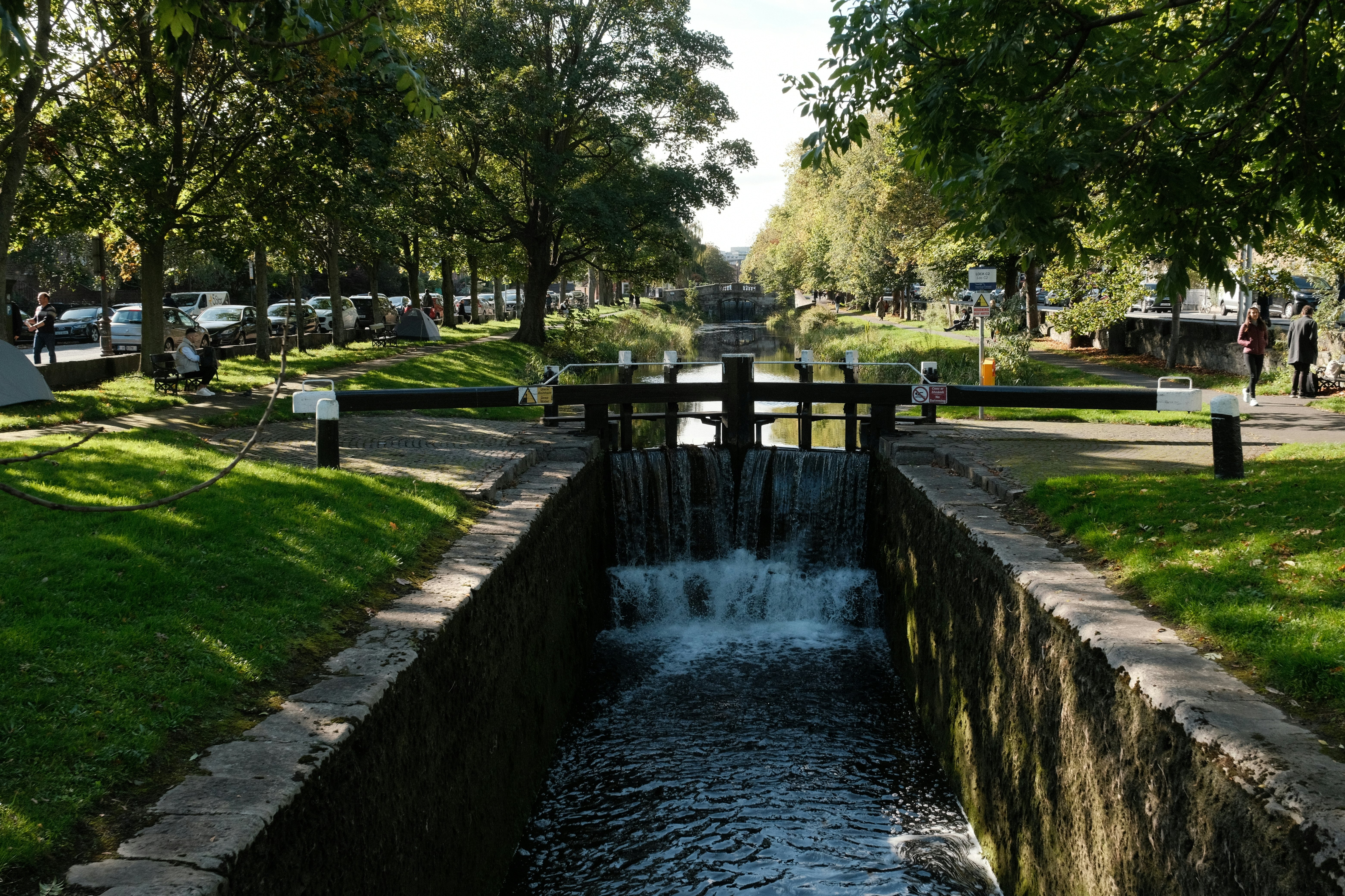 A small lock in a narrow canal lined by grassy banks in a city.