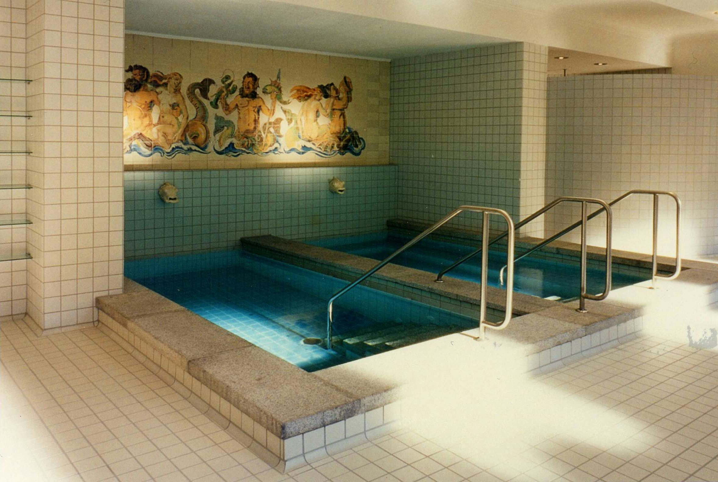 Steps lead down into two small rectangular indoor pools. A mosaic of people at sea is on the wall above.