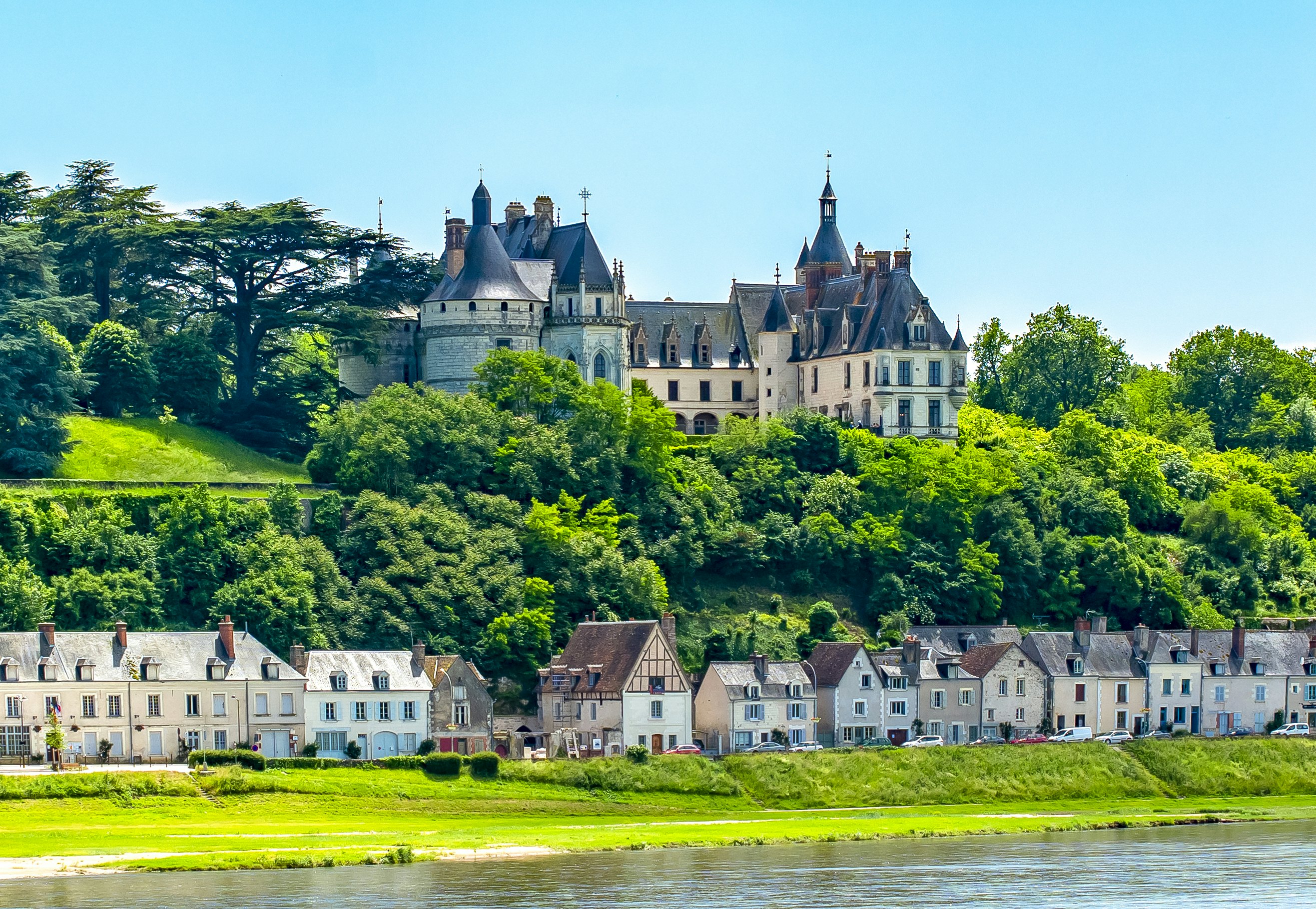 Houses line the green banks of a riverside overlooked by a large chateau.