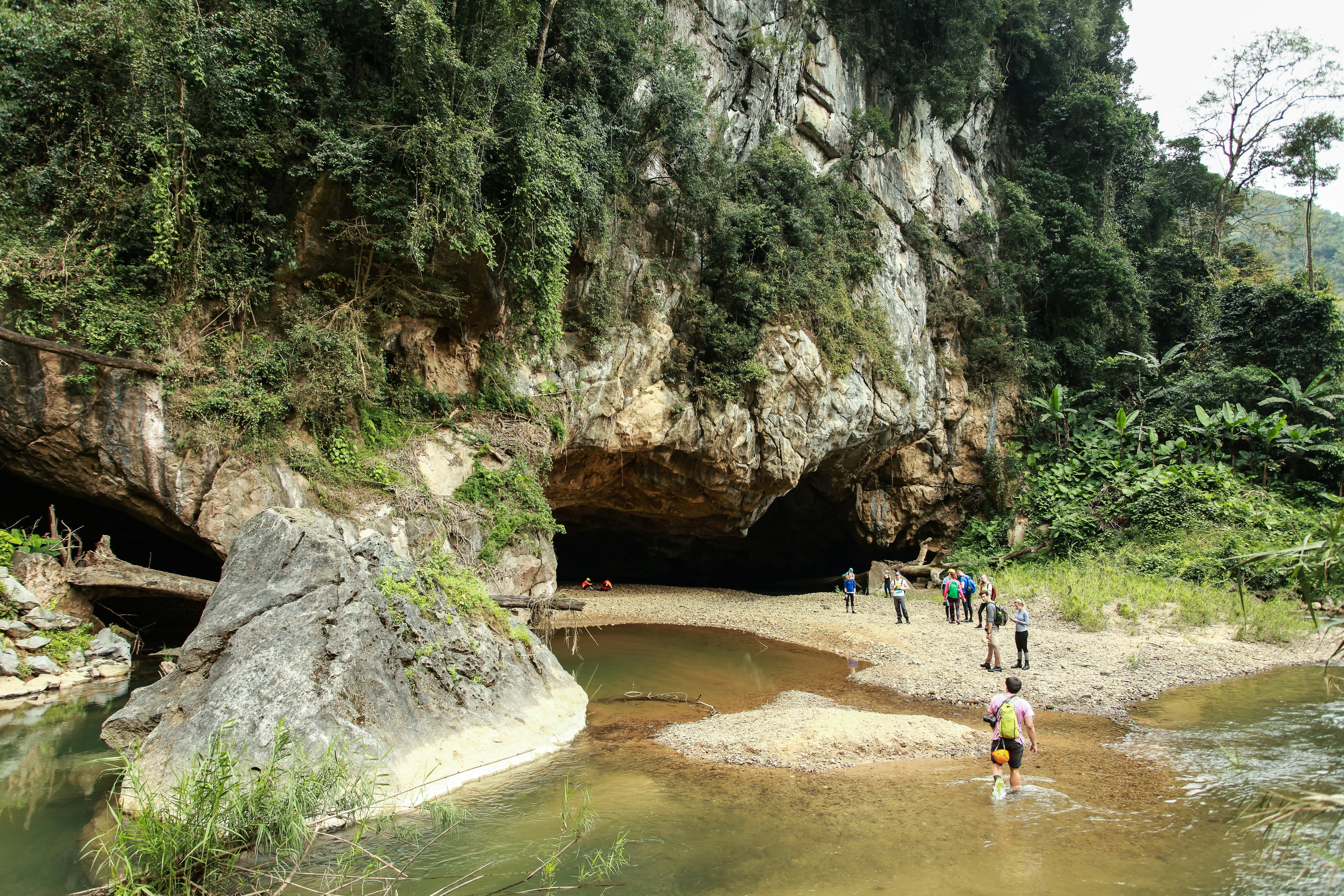 People wade through a river on their approach to a cave mouth surrounded by jungle.