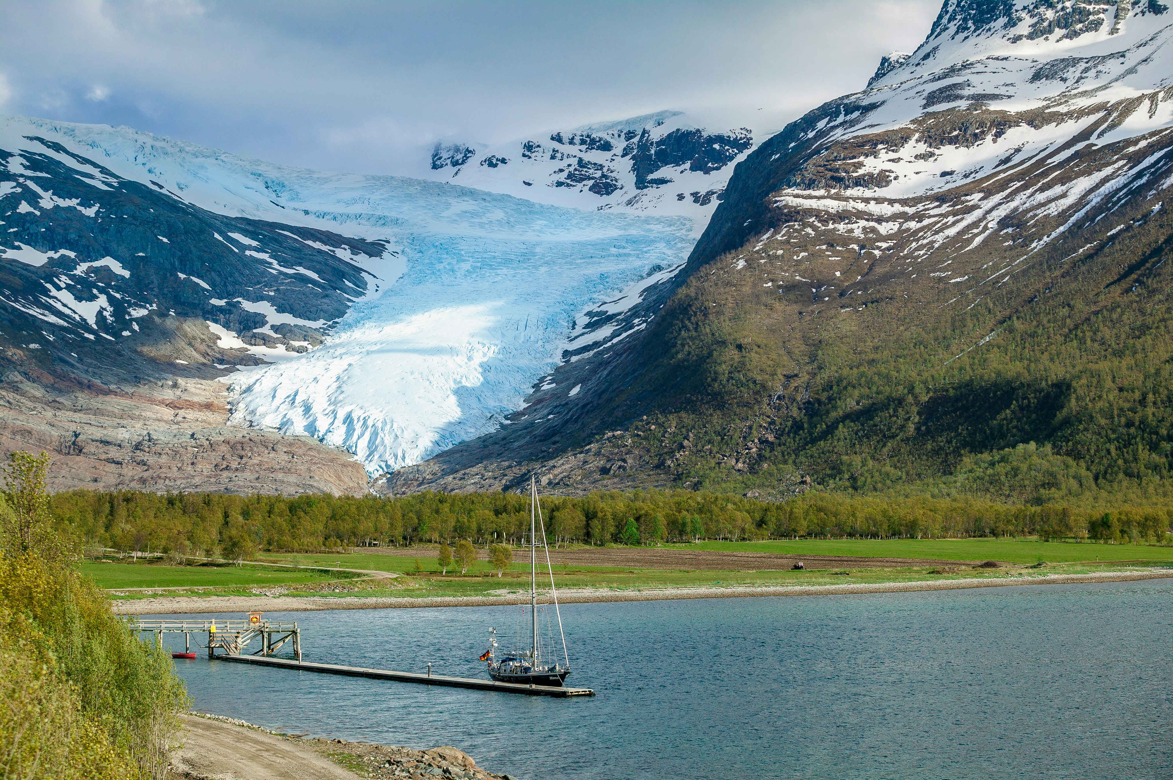 A sailing yacht with sails down, moored at a dock with a mountain and glacier in the distance on a sunny day.