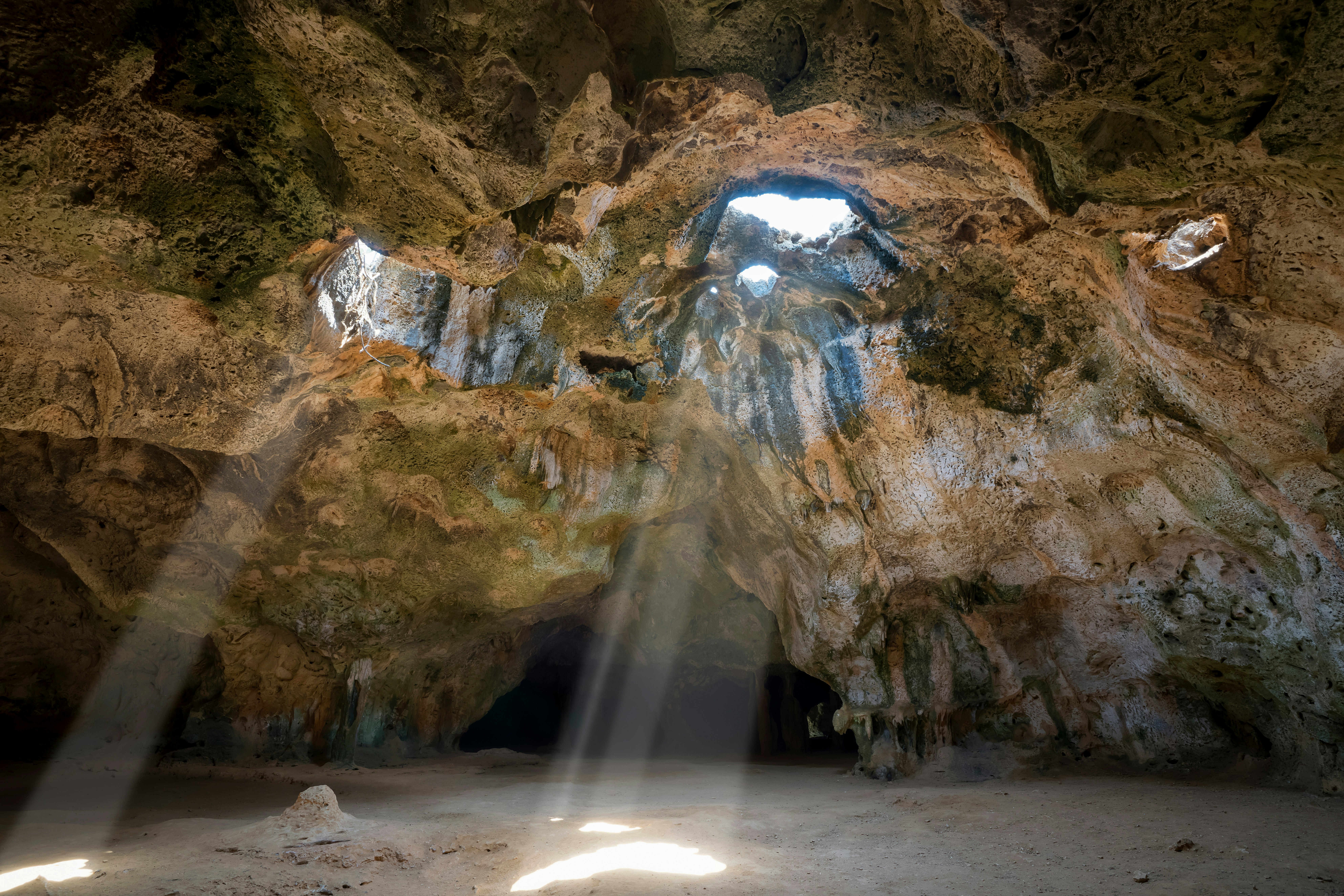 Light streams through openings in a rock cave in Aruba to shine on white sand.