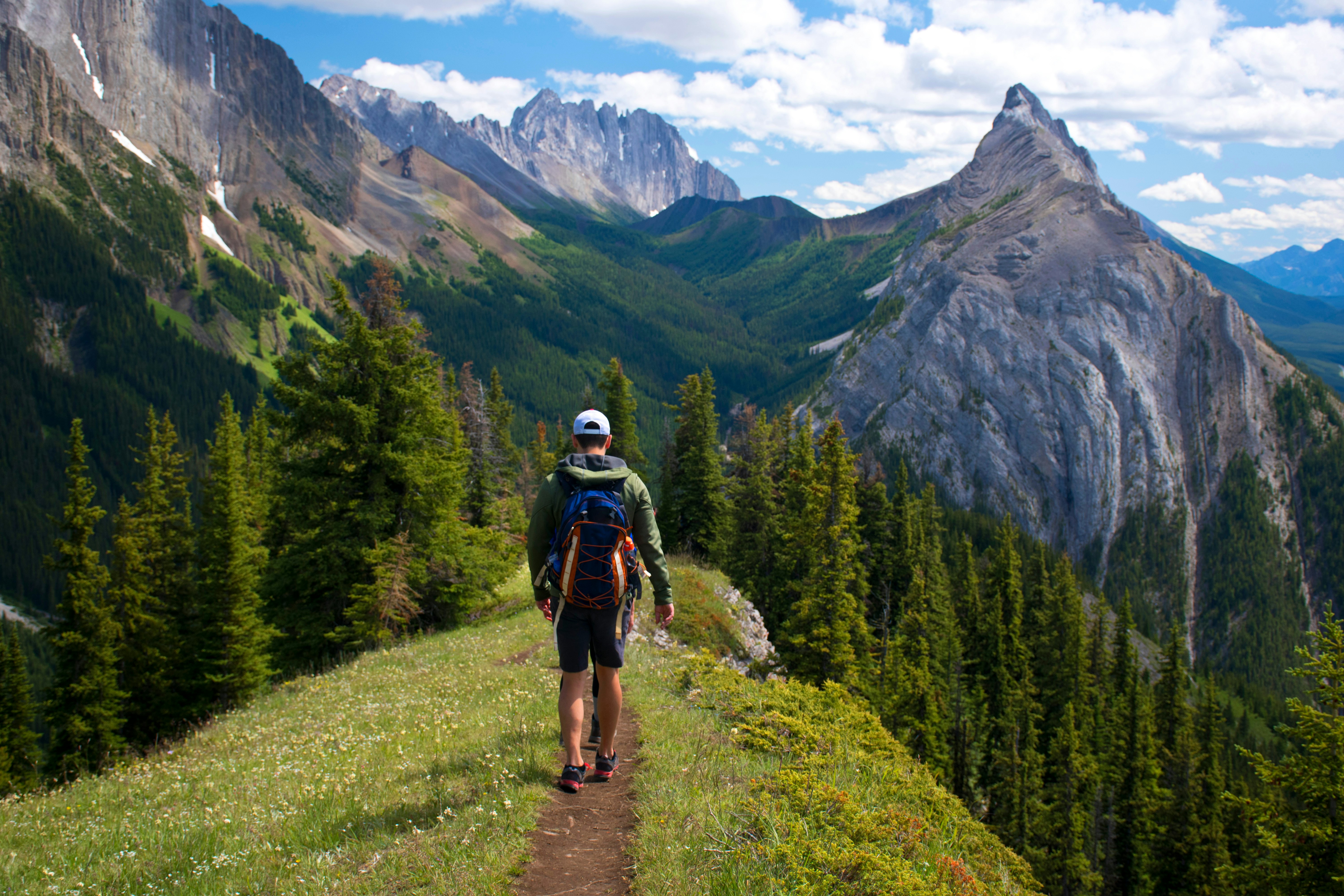 A man wearing shorts and a backpack walks on a narrow dirt path on a ridge covered with grass and flowers heading toward steep rocky peaks.