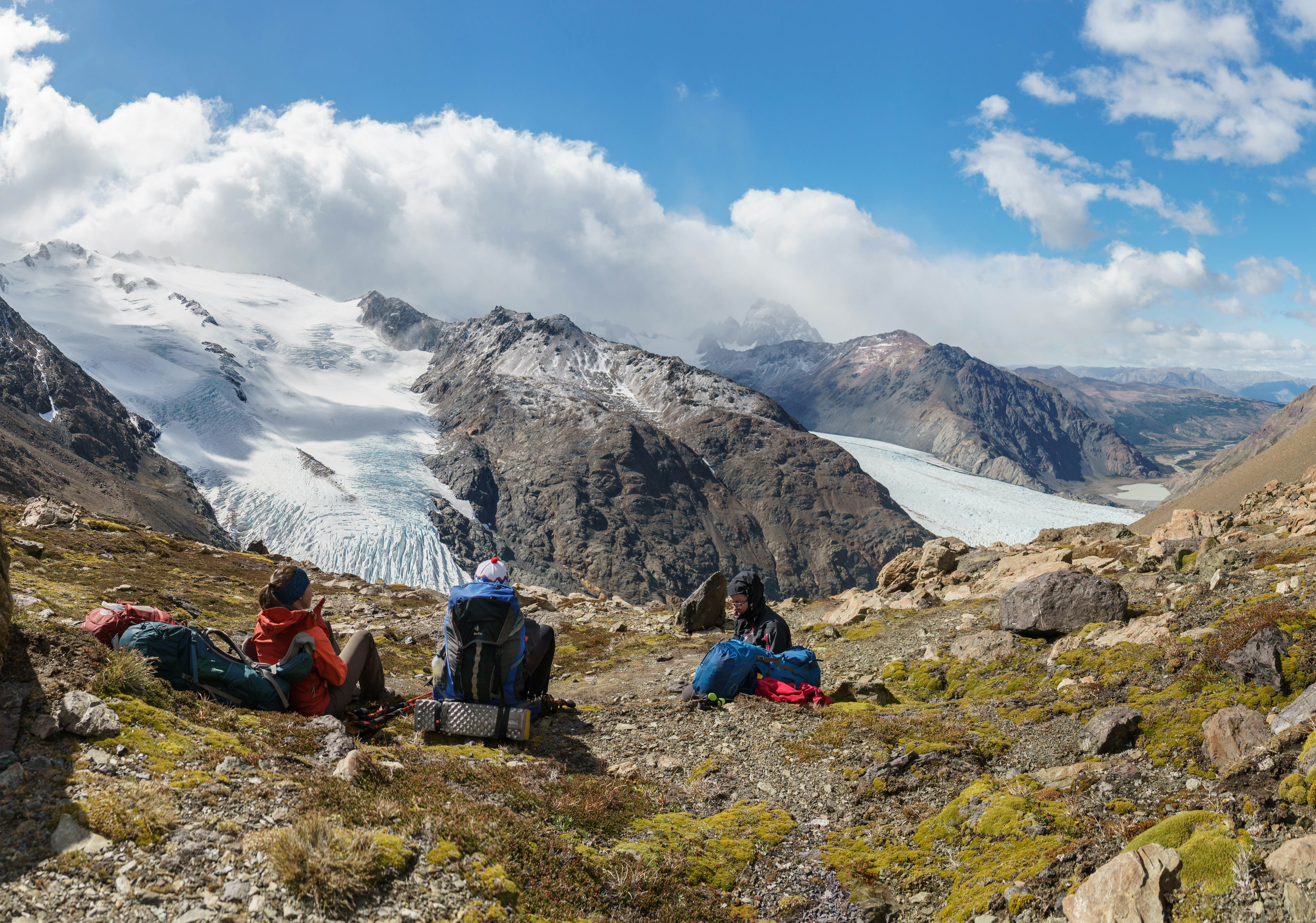 Three hikers sit on a patch of grass looking at tips of glaciers extending down a rocky mountain.