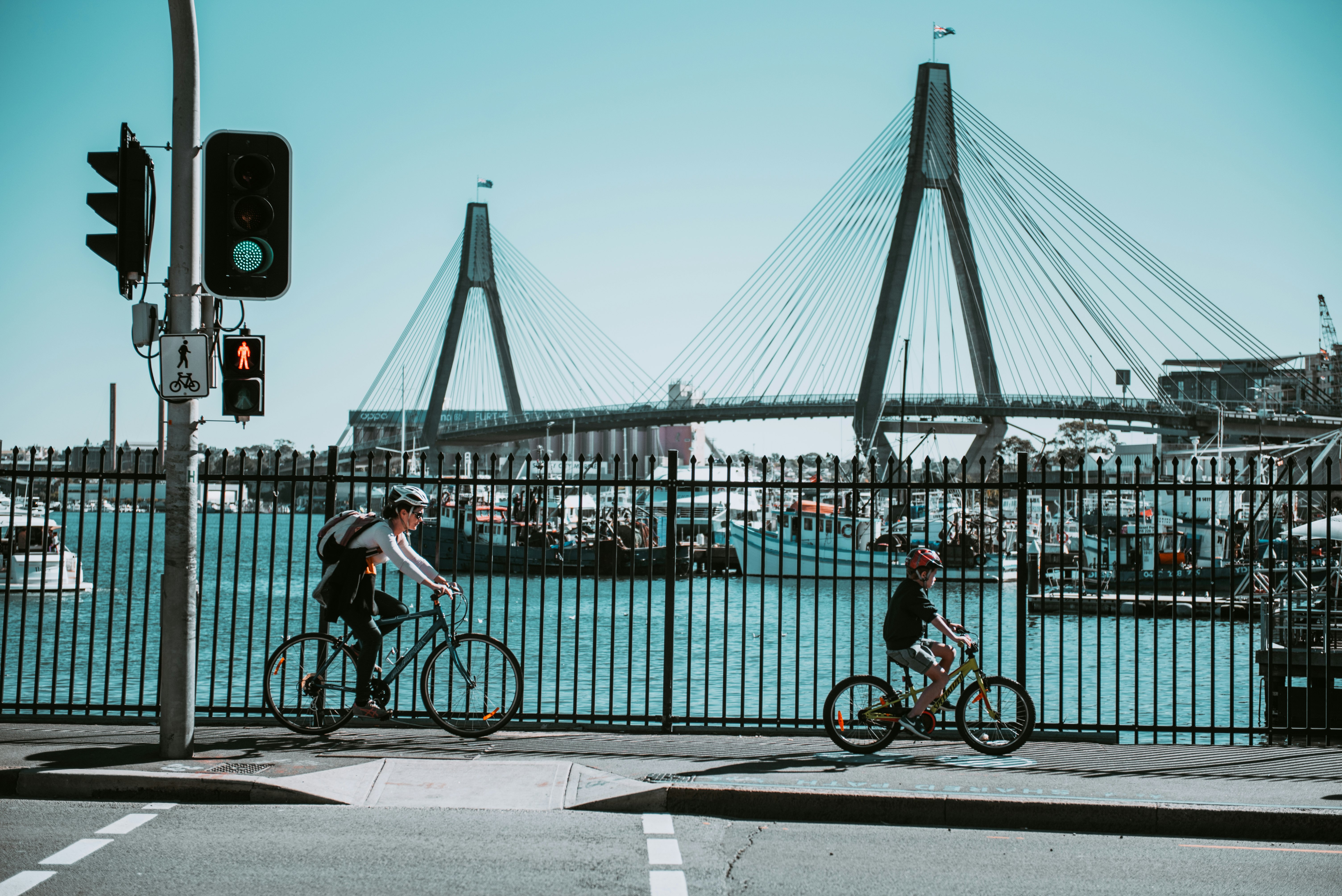 A woman and a child ride bikes on the sidewalk near a harbor. A large suspension bridge passes behind them.