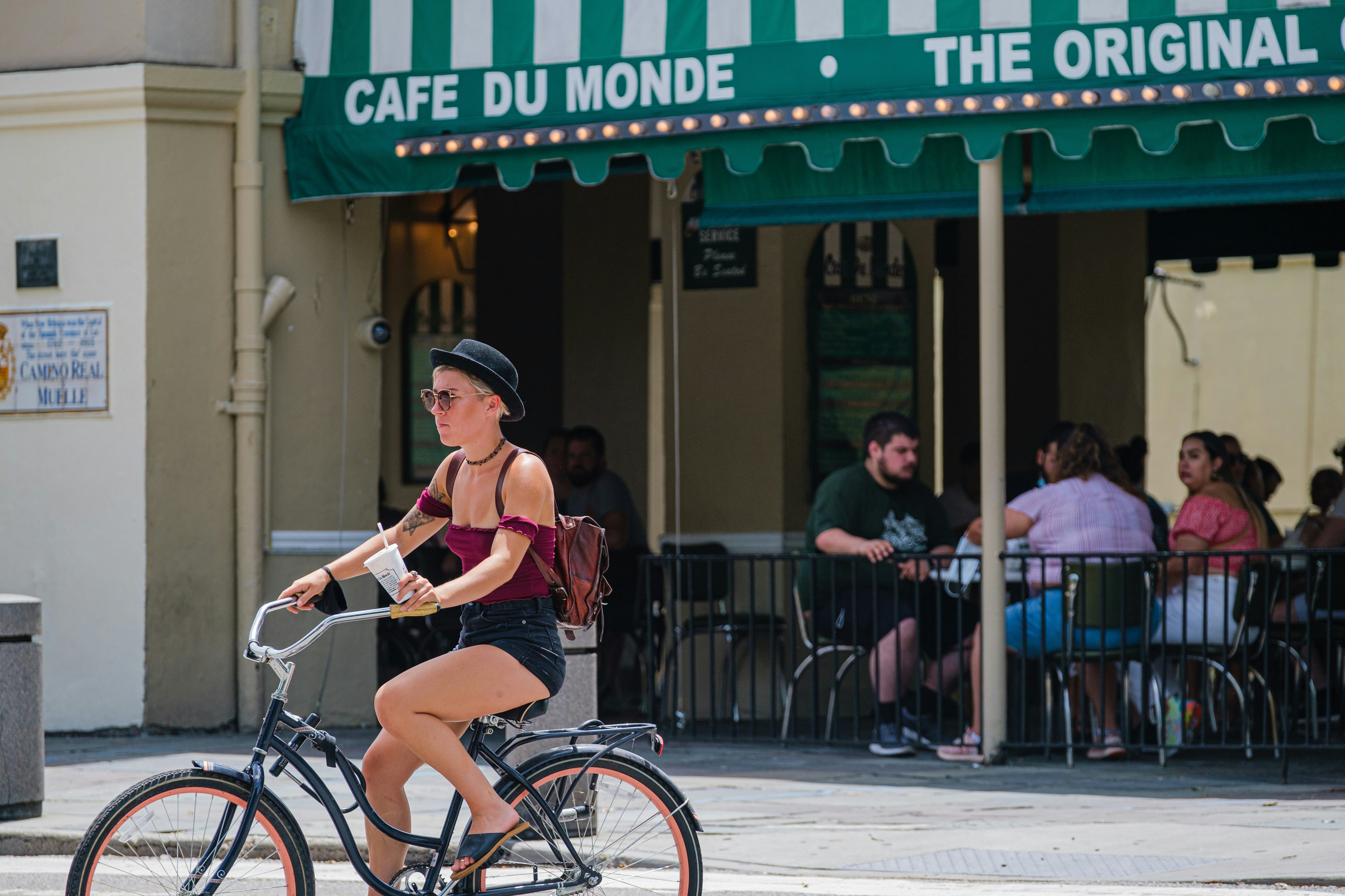 A female cyclist in a black hat pedals past a cafe with outdoor tables under a green awning.