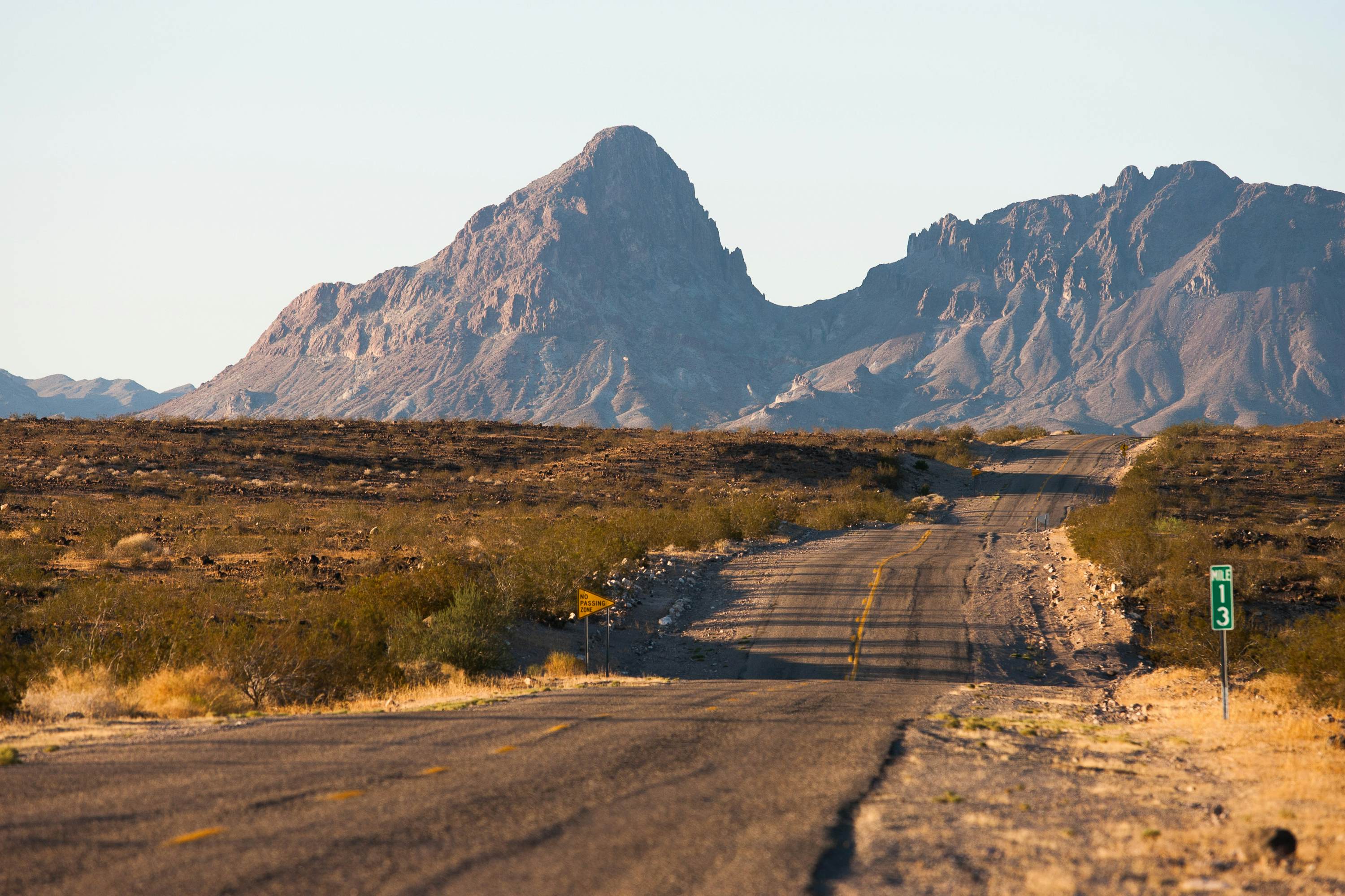 A two-lane road winds through low green bush with large rocky mountains in the background.
