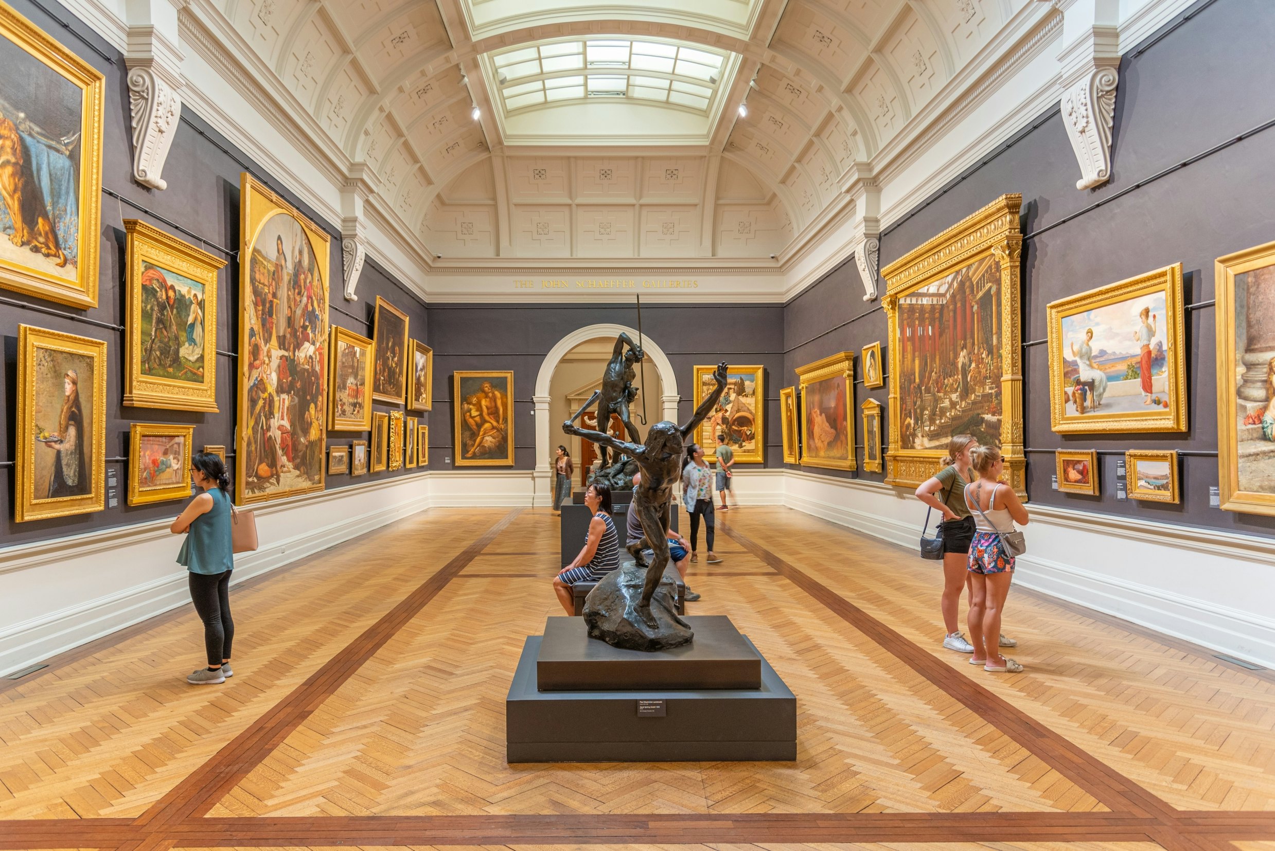 An art gallery hall with walls lined with works of various sizes. Sculptures are in the middle of the hall.