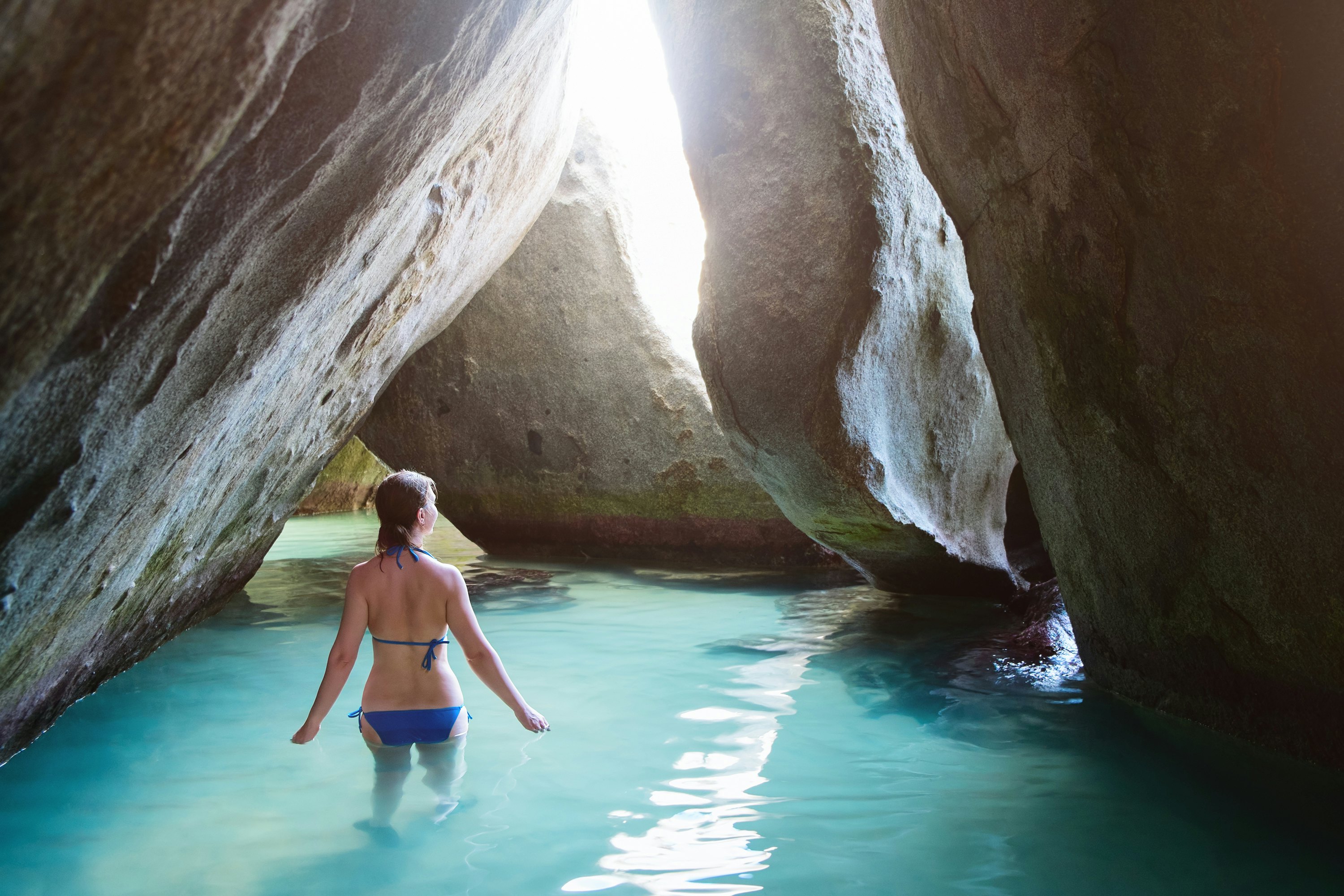 A woman wades in water in a cavern formed by boulders. Sunlight streams in through an opening in the large rocks.