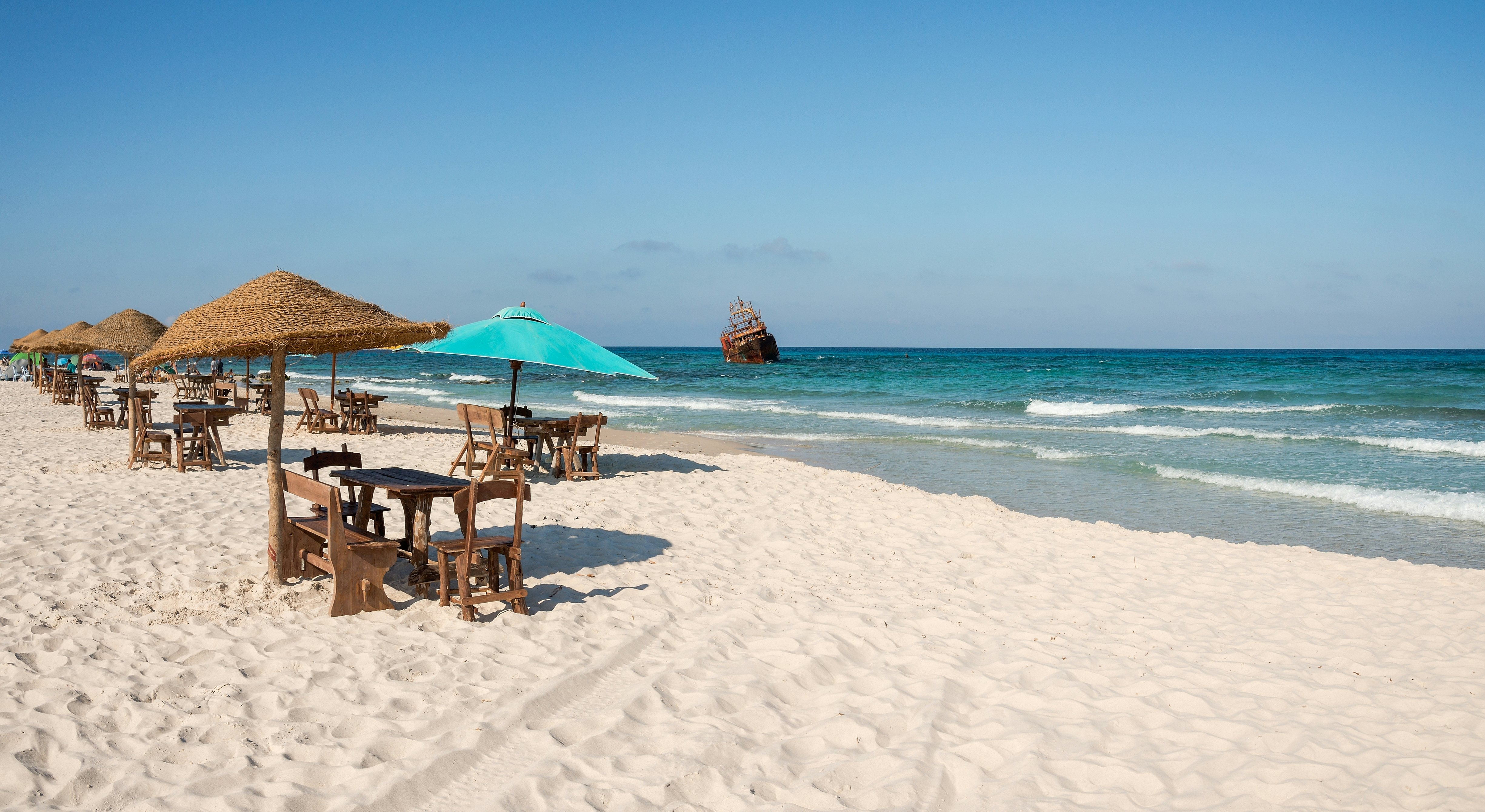 Tables and straw umbrellas are seen on a white-sand beach. The rusted hulk of a ship sits in the shallow water offshore.
