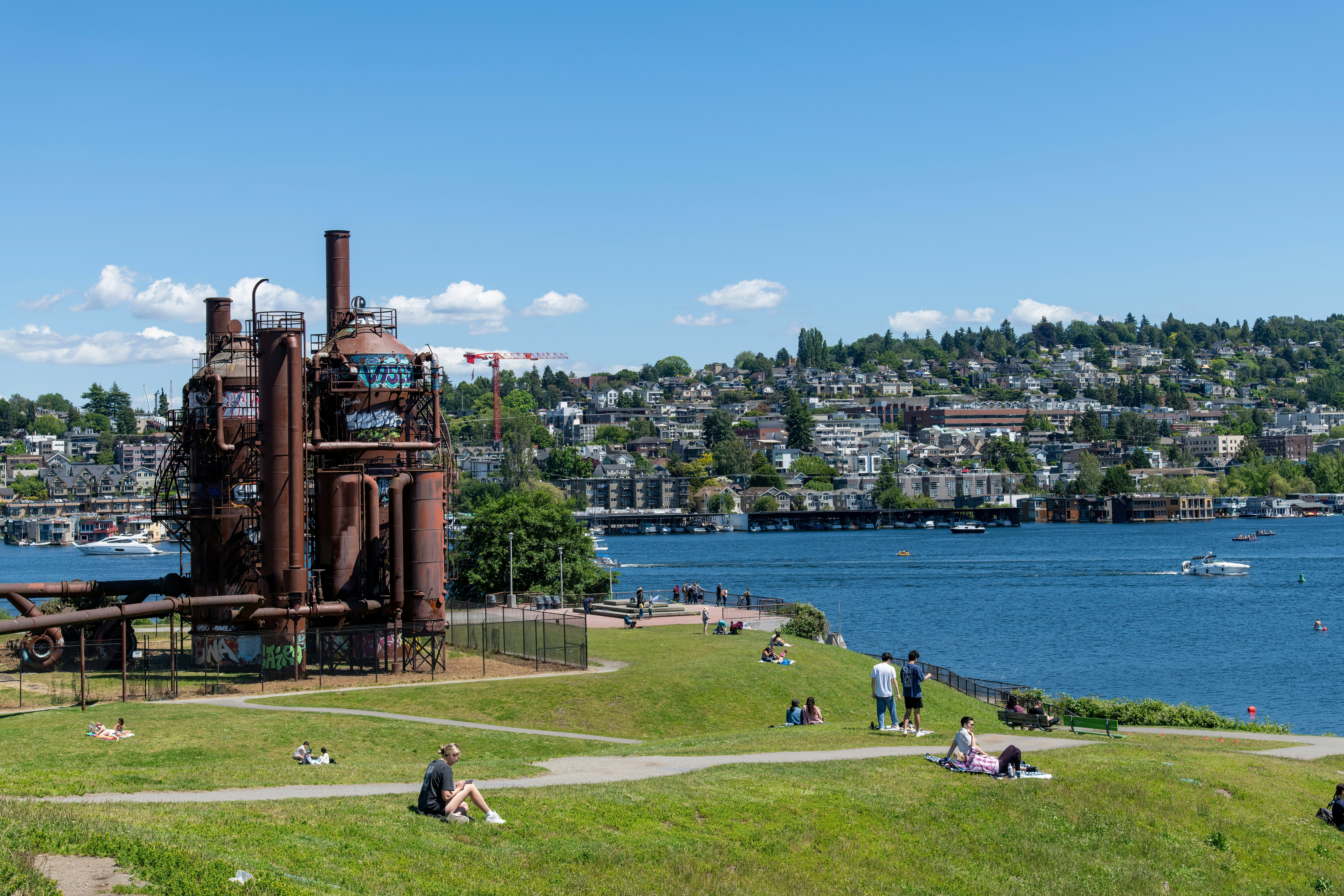 People sitting on the grass on the lakeshore at a park. Buildings and trees rise from the hill across the lake.