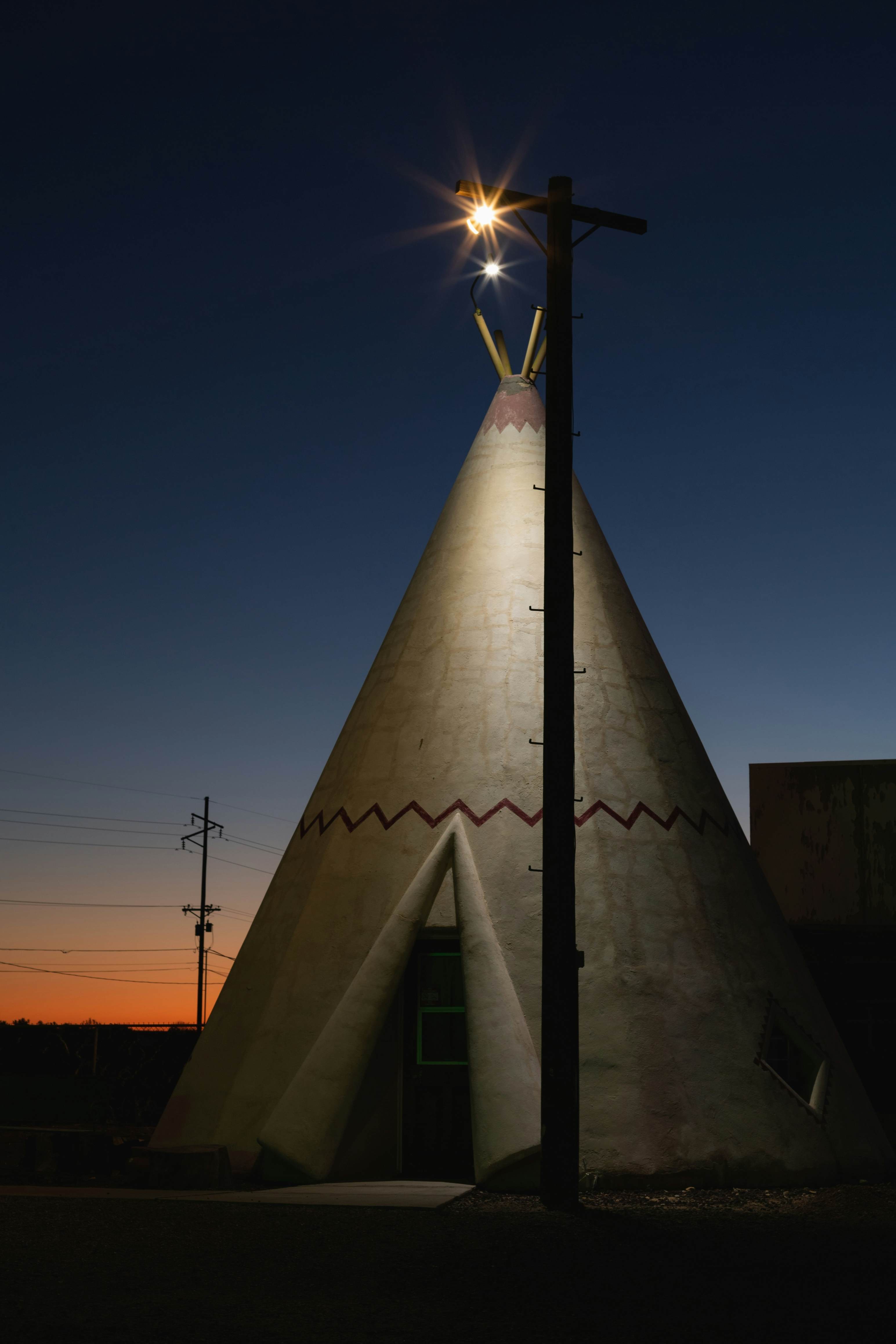 A pink, orange and deep blue sunset behind a teepee illuminated by a single street light