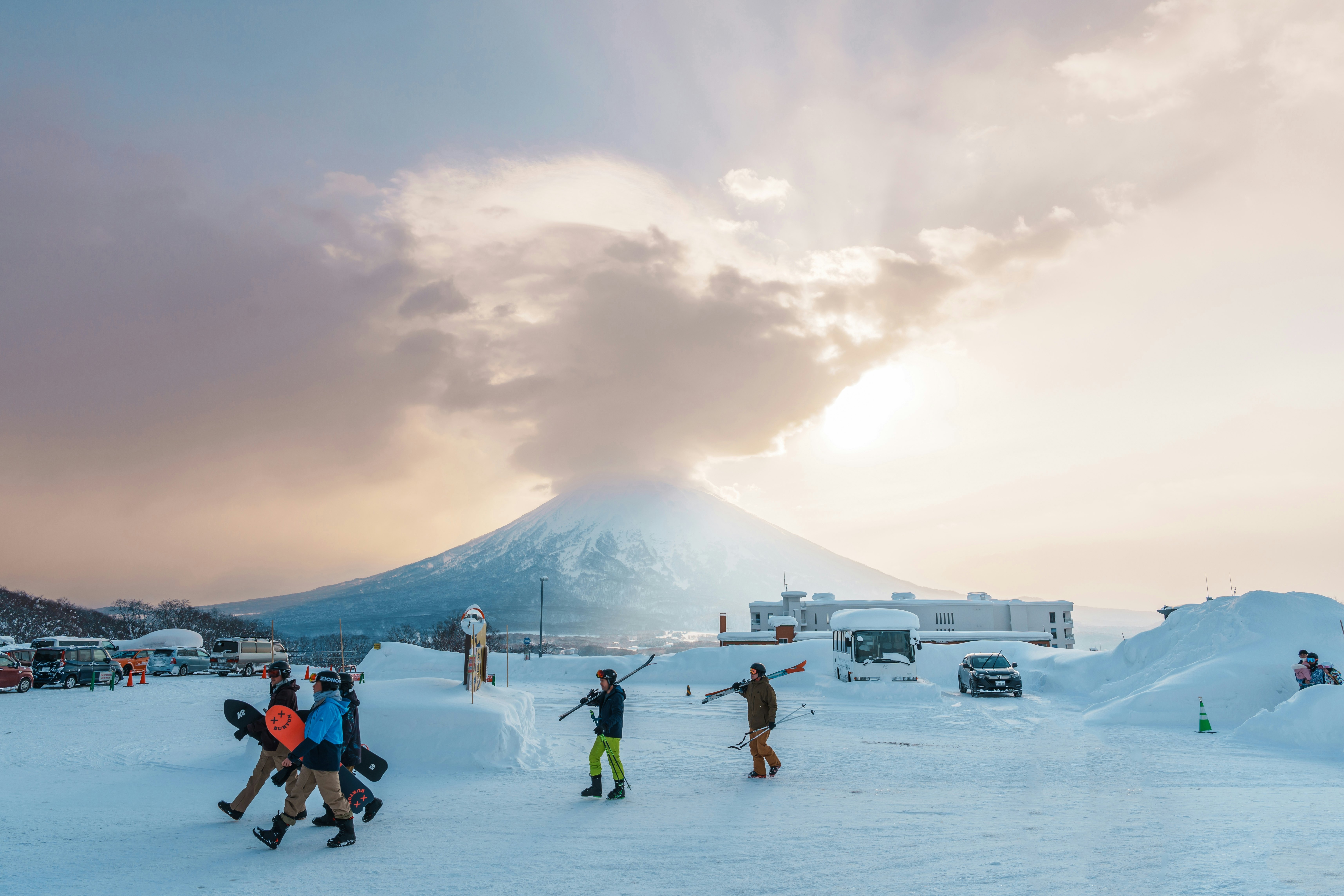 People carry their skis and snowboards at a snowy mountain resort. A huge stratovolcano is visible in the distance.