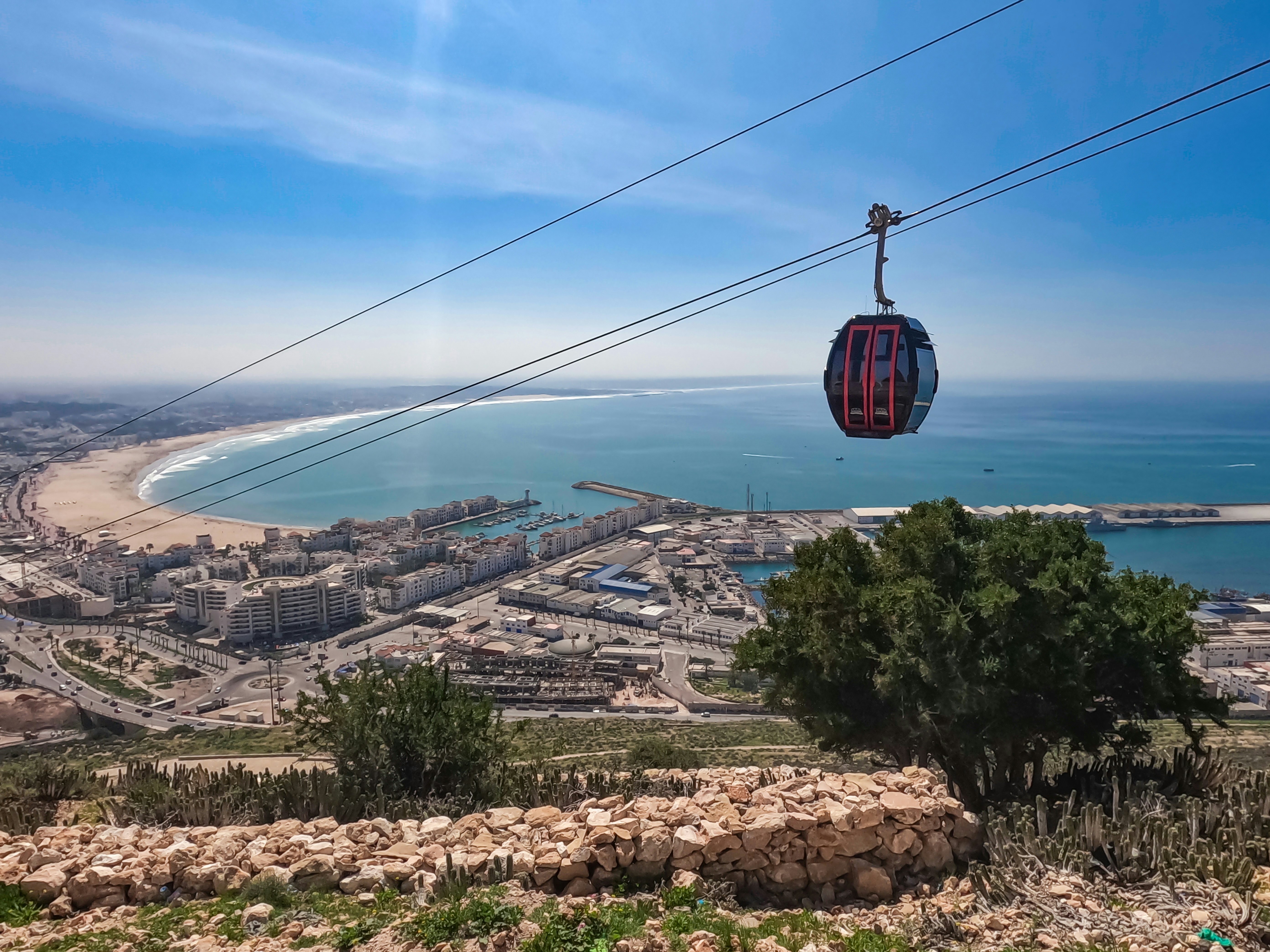A cable car suspended over a hillside above a port city; buildings and a wide beach border blue water.