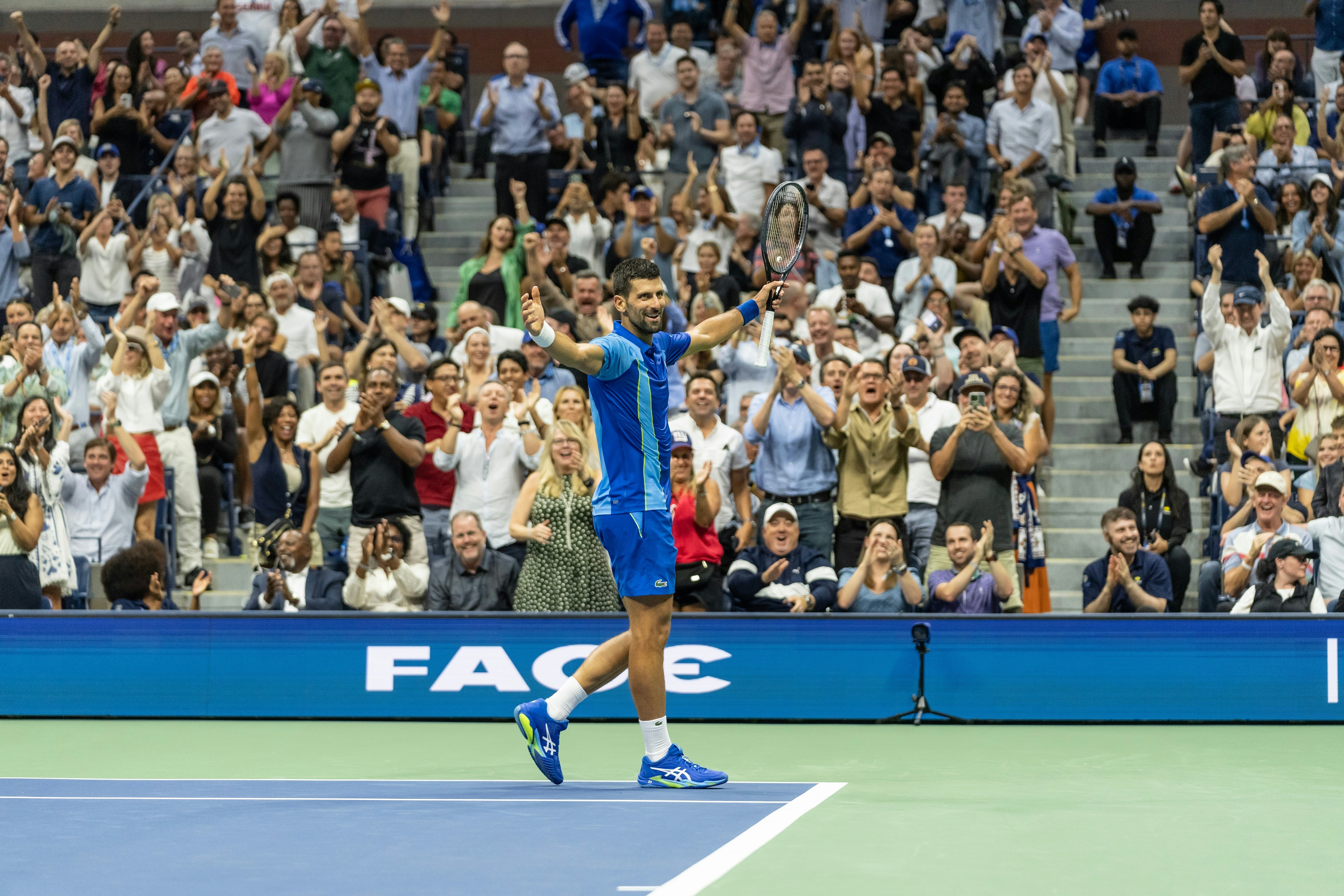 A male tennis player walks off the court with his arms outstrethce smiling as a crowd of people beyind him stand and cheer.