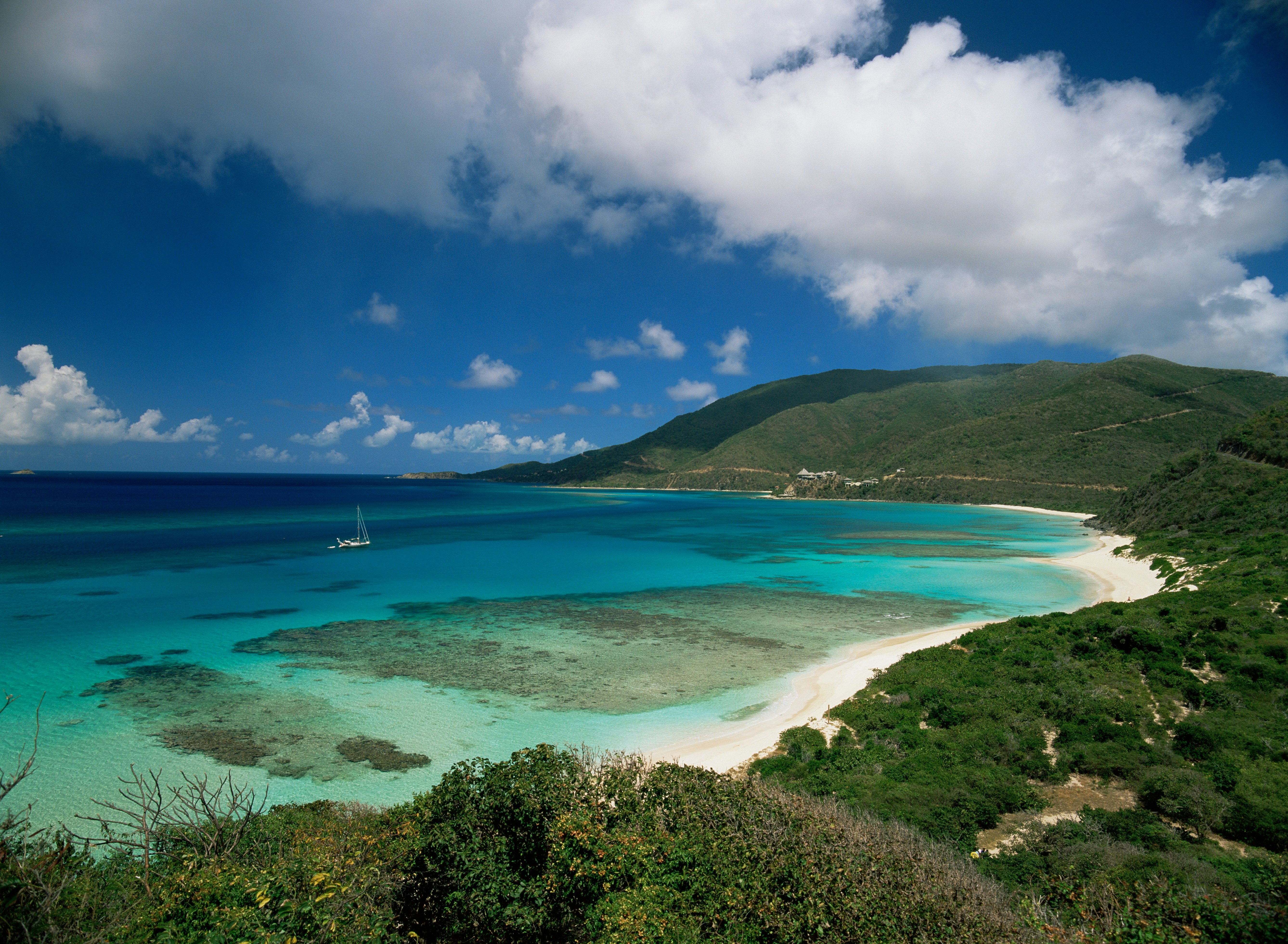 A wide view of a cove and beach on a tropical island. The waters are many shades of blue, and the vegetation on land very green and lush.