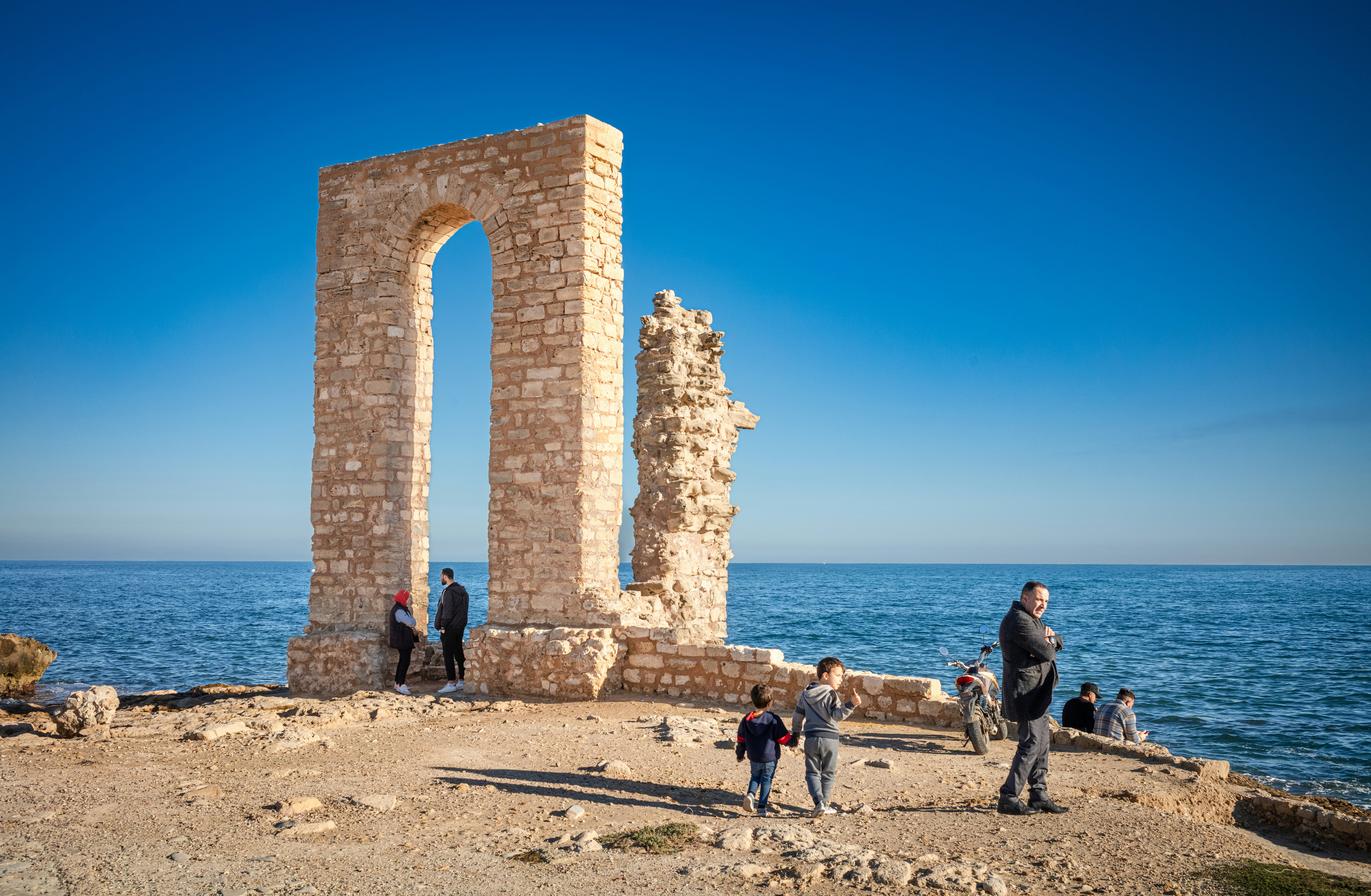 People walk near a reconstructed stone arch that rises on rocky ground next to the sea.