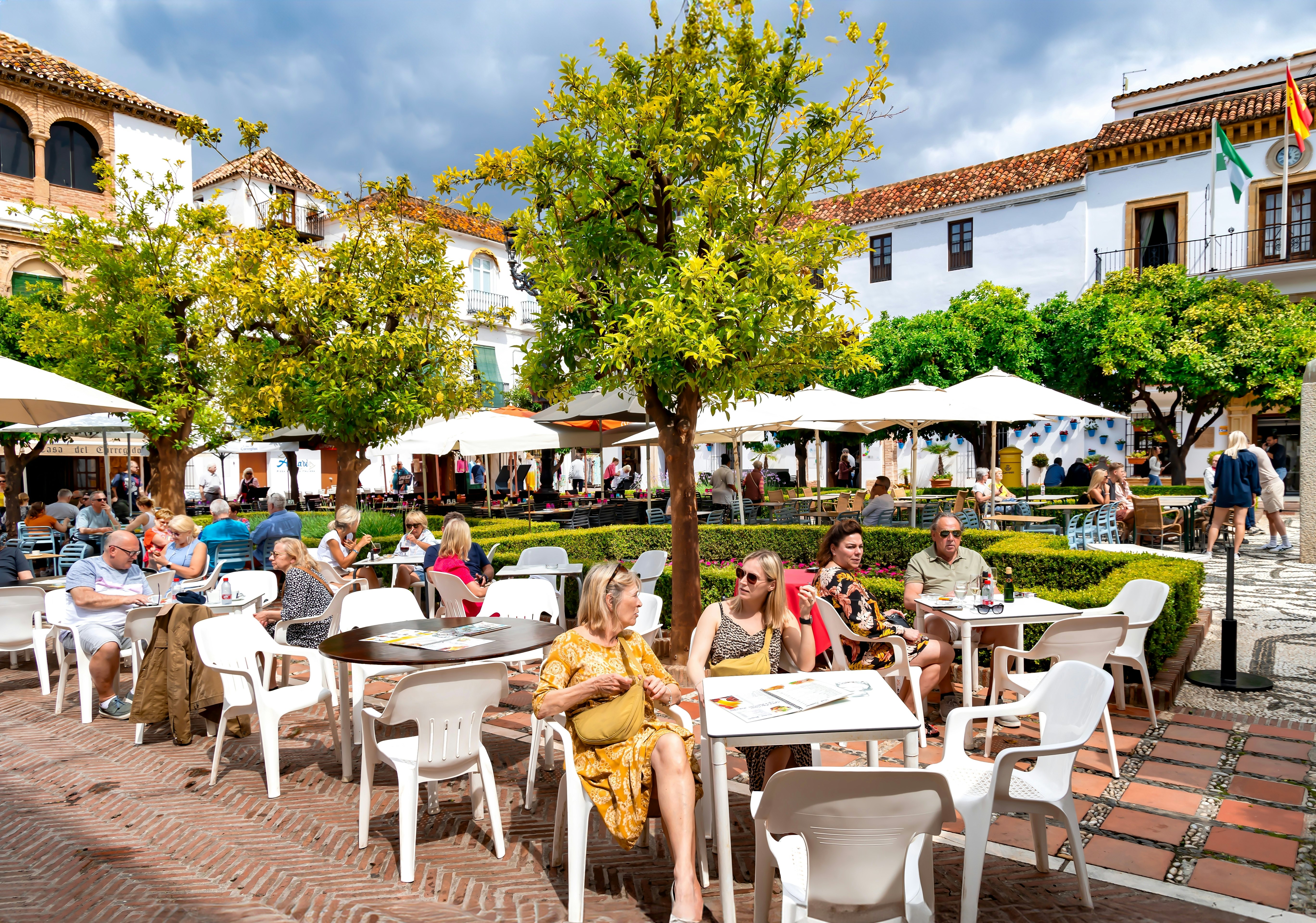 On a sunny day in Spain, people sit under orange trees at white tables arranged around a central square that has low hedges.