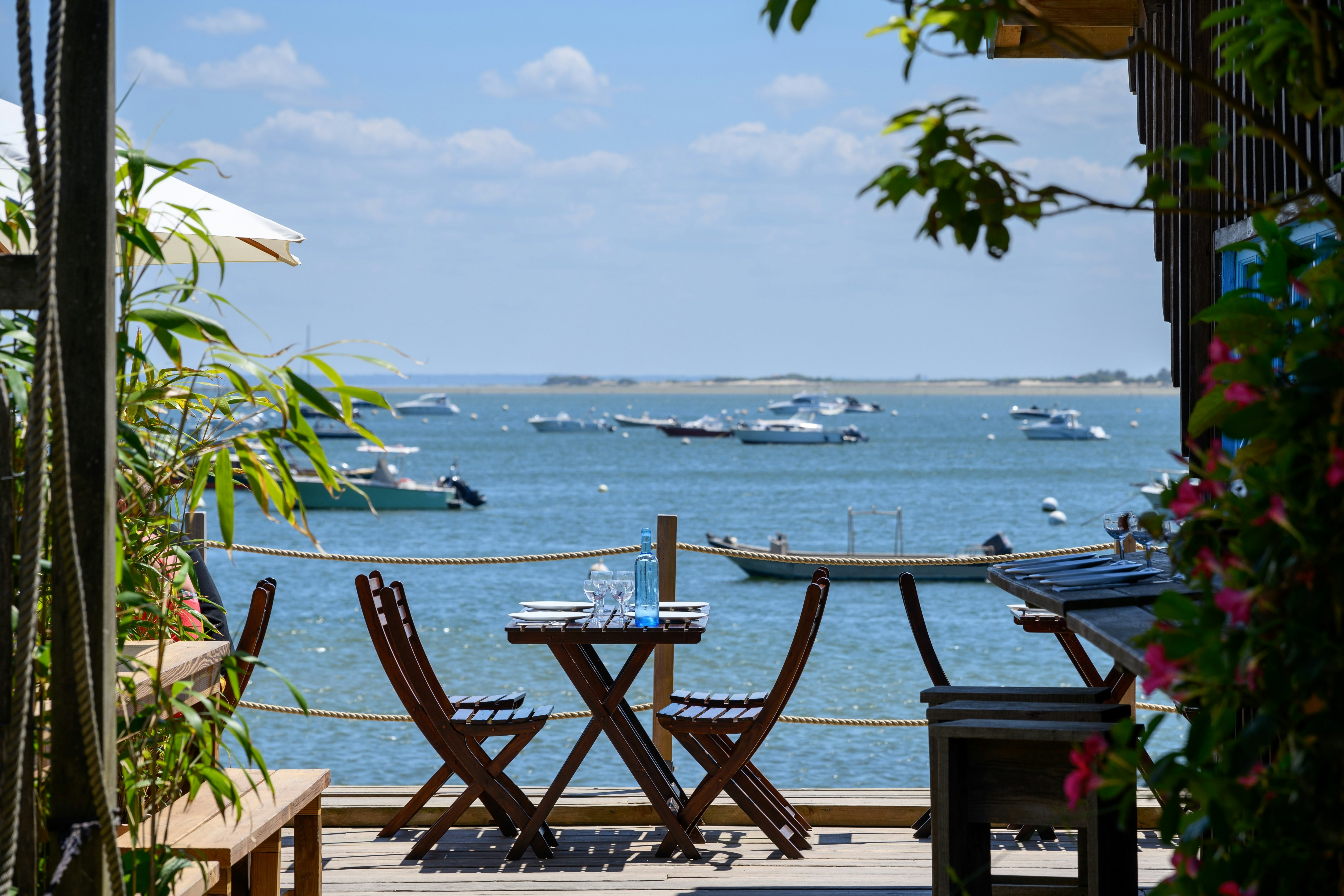 A table set on the decking of a casual seaside restaurant. Boats are docked in the bay.