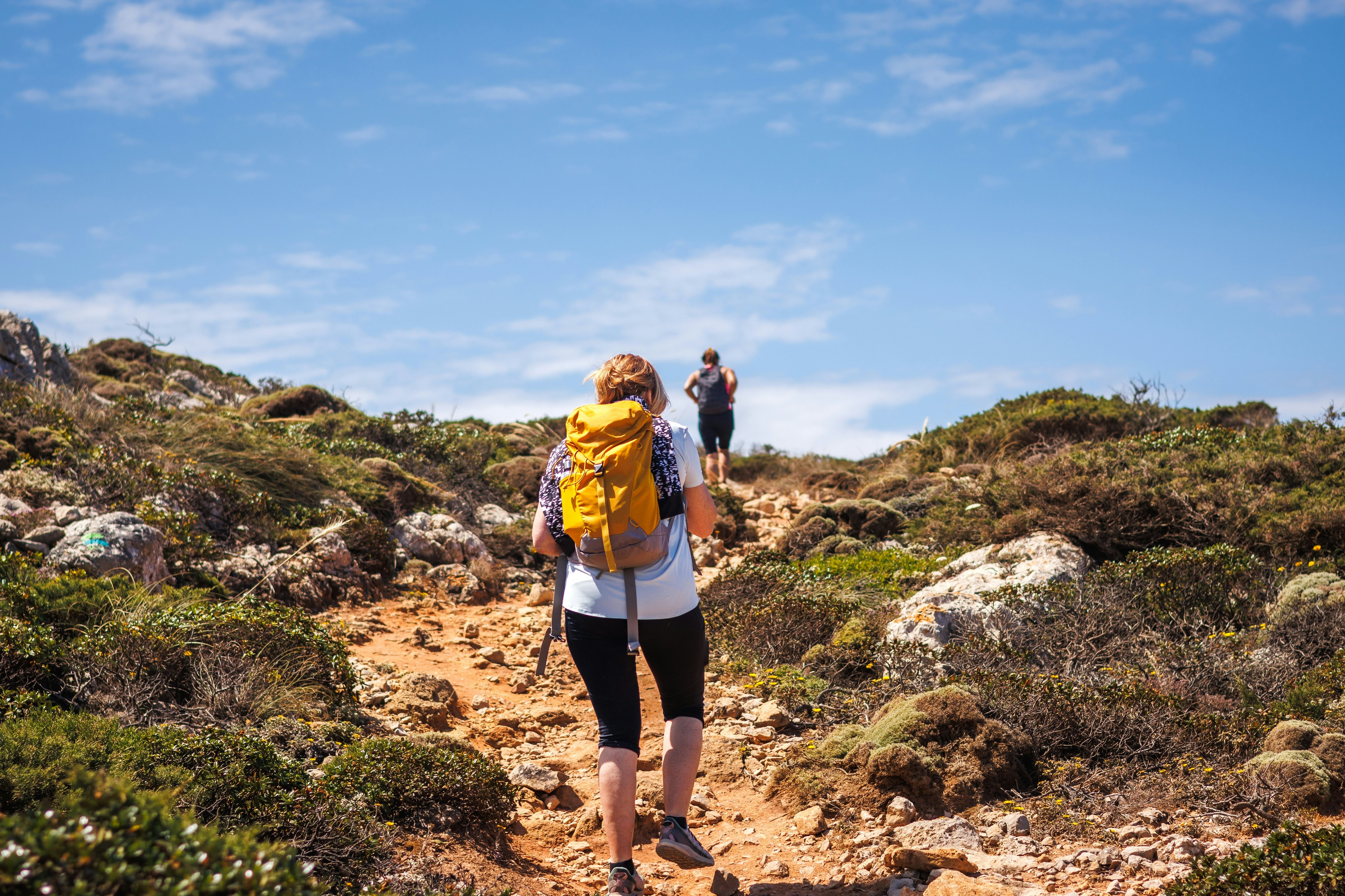 Two hikers with backpacks walk on a rocky path up to cliffs.