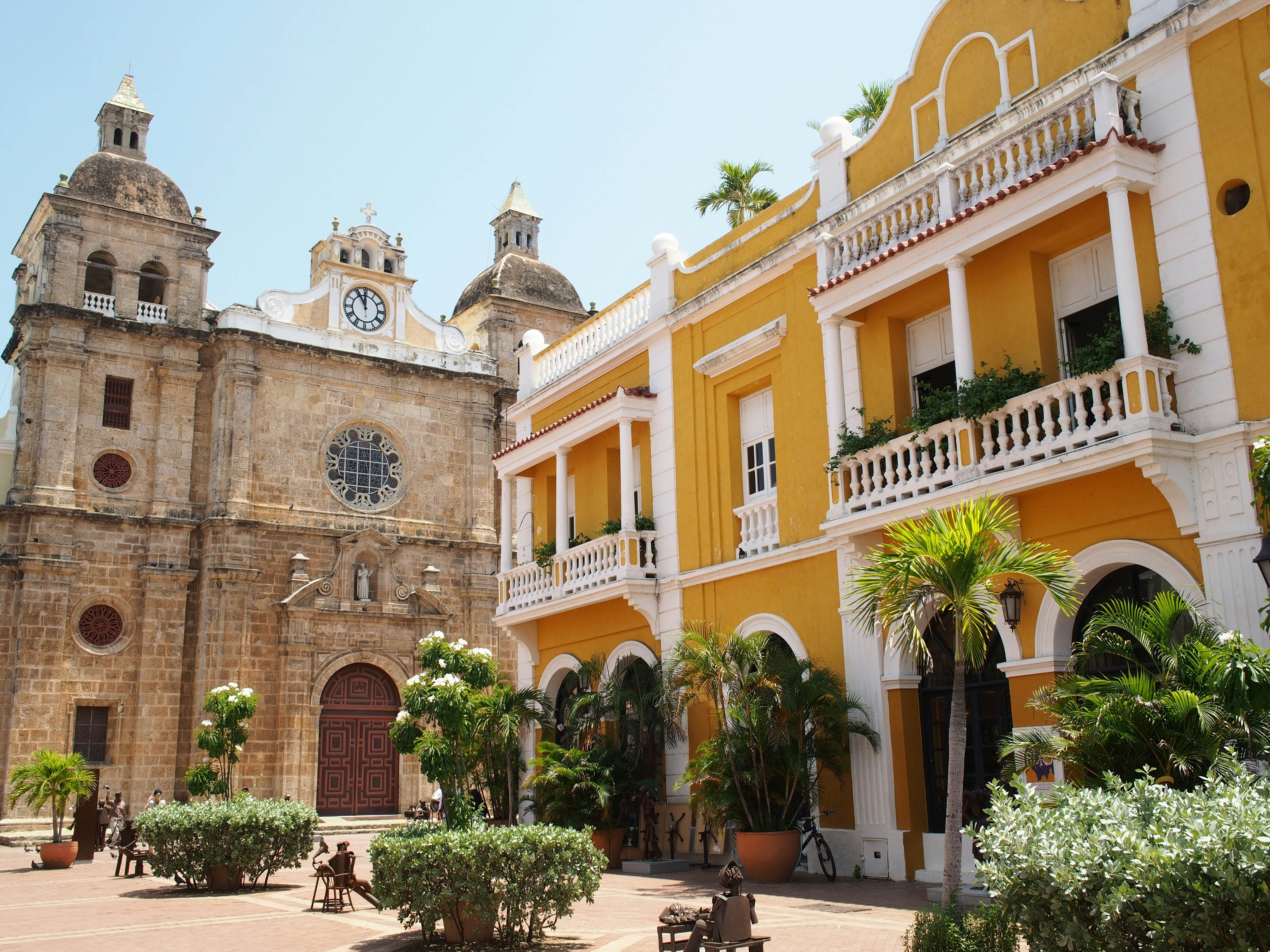 A tan stone church and a yellow-and-white building are positioned at one end of a plaza with tropical plants in the center.