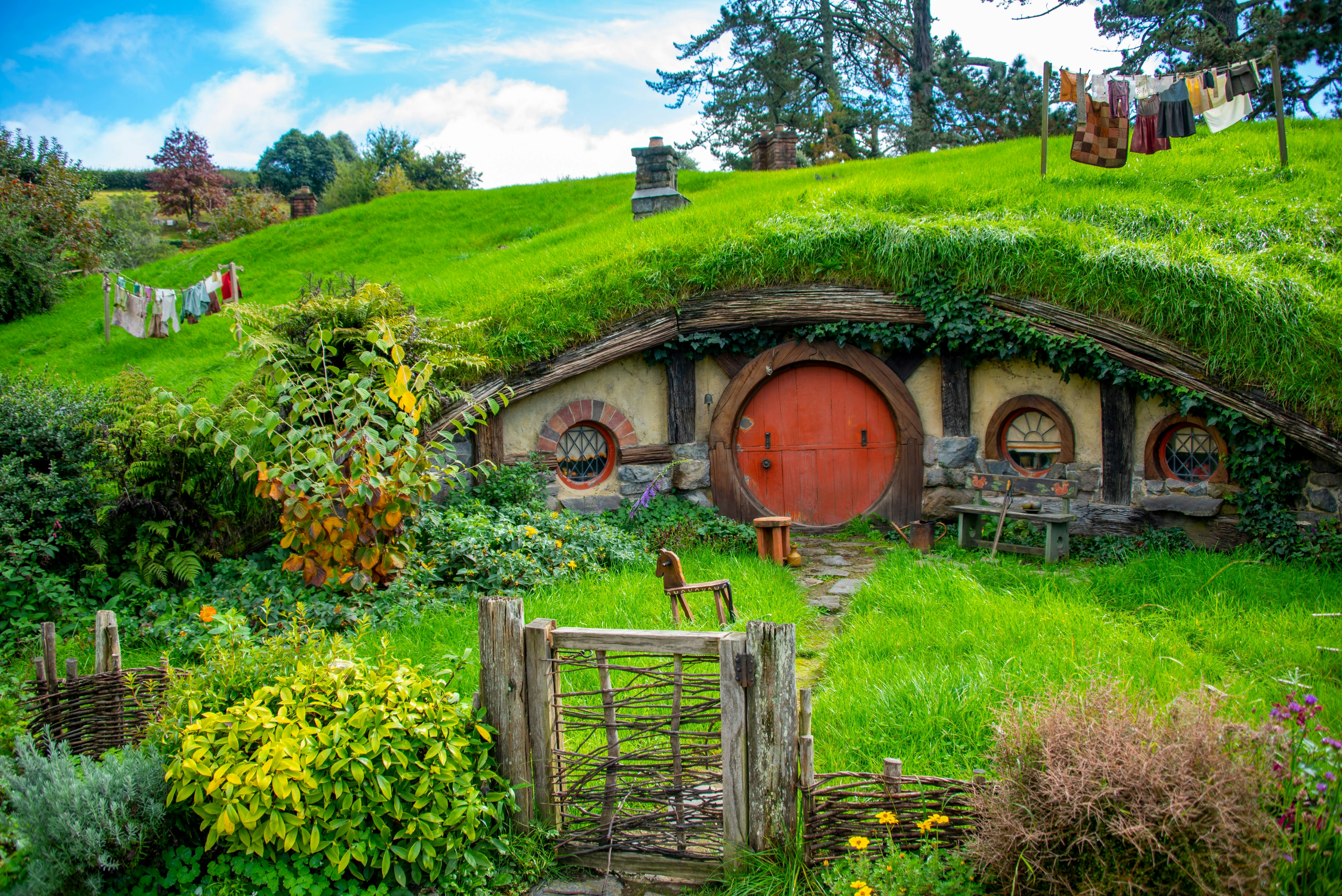 A small curved cottage set into a hillside of bright green grass with a rustic gate in front and a red door on the cottag on a sunny day.
