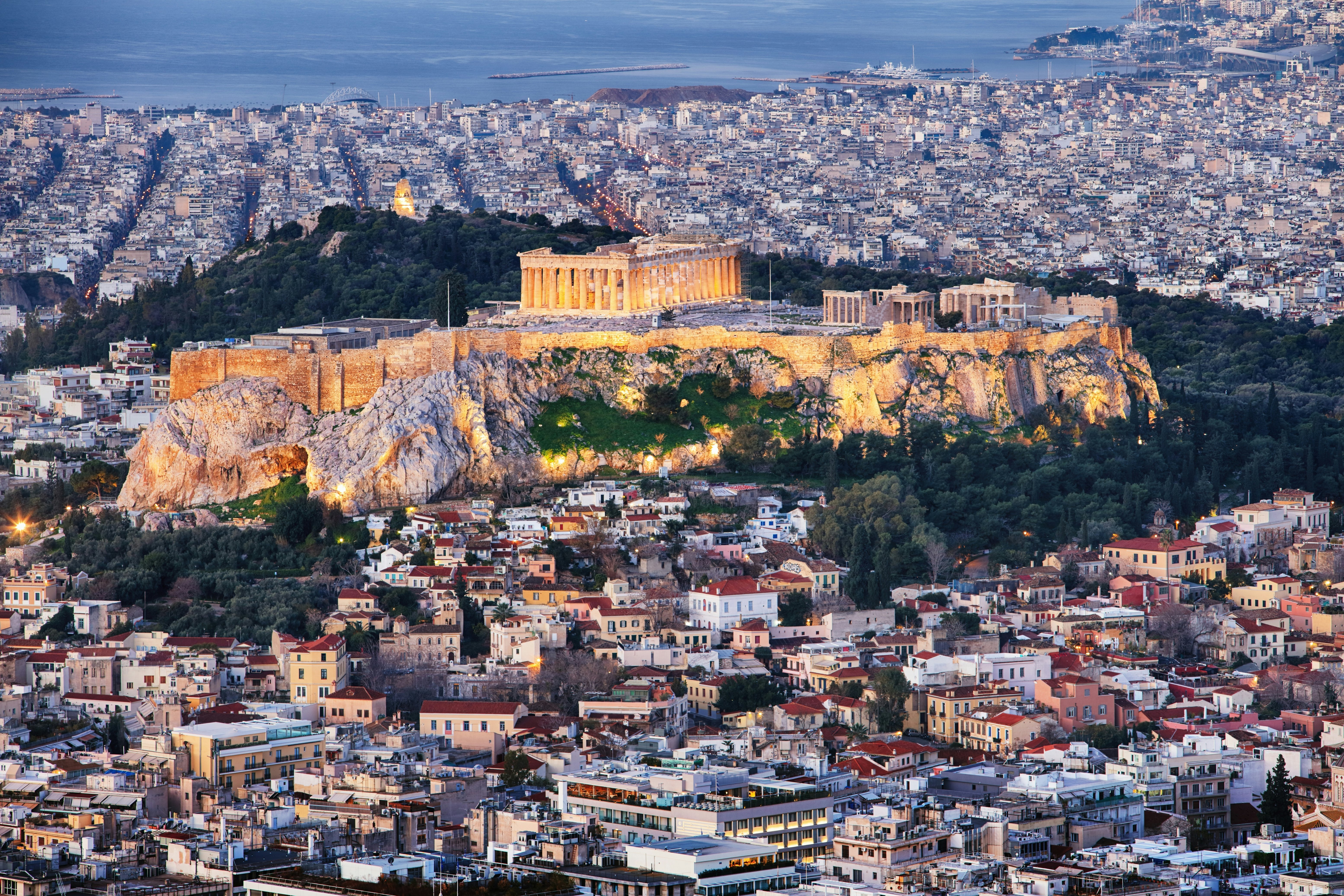 Aerial view of ruins on a hillside with a city surrounding it and the ocean beyond.