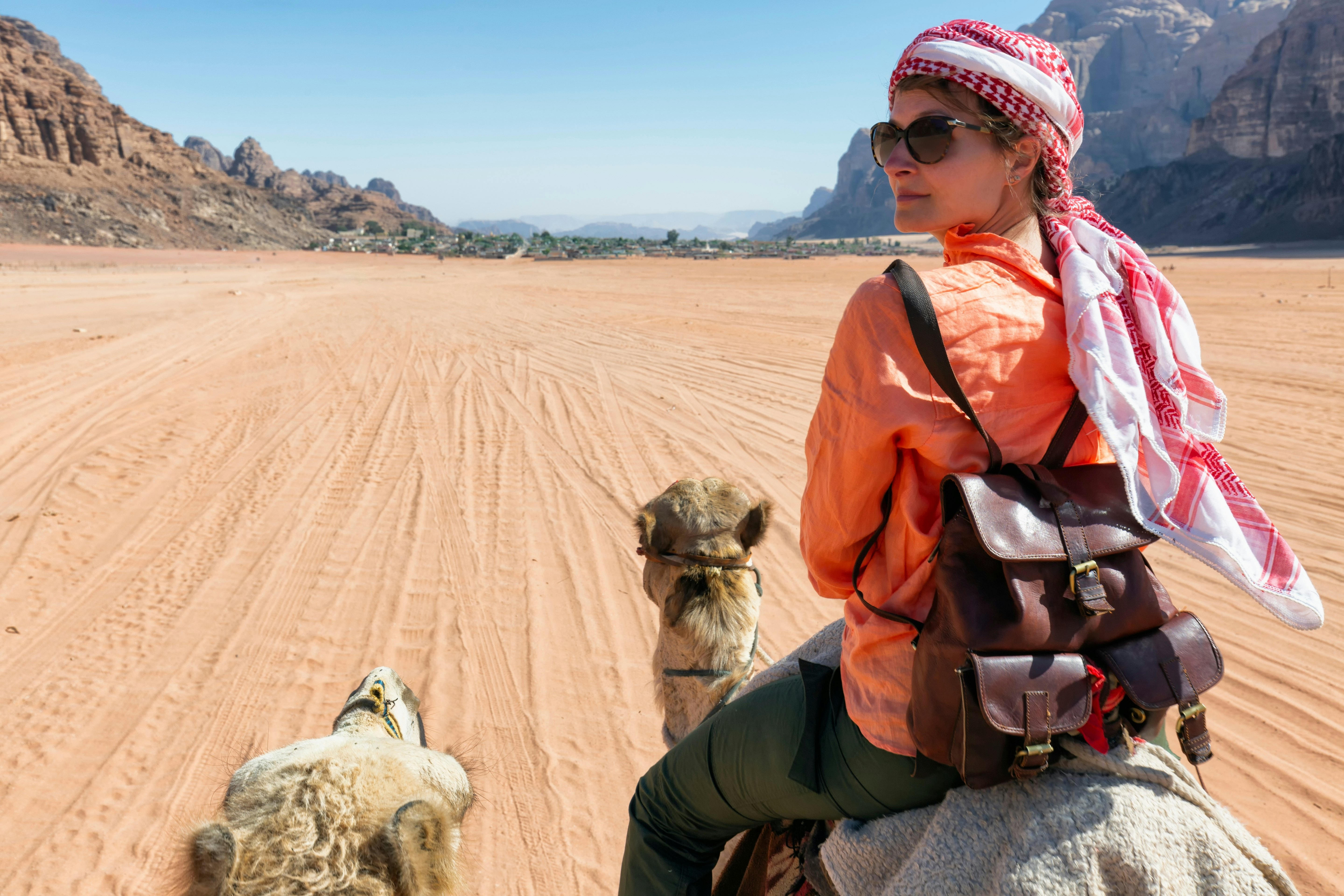 A person riding a camel wearing a head wrap looks over their left shoulder; the landscape ahead is reddish sand with rock formations on the perimeter.
