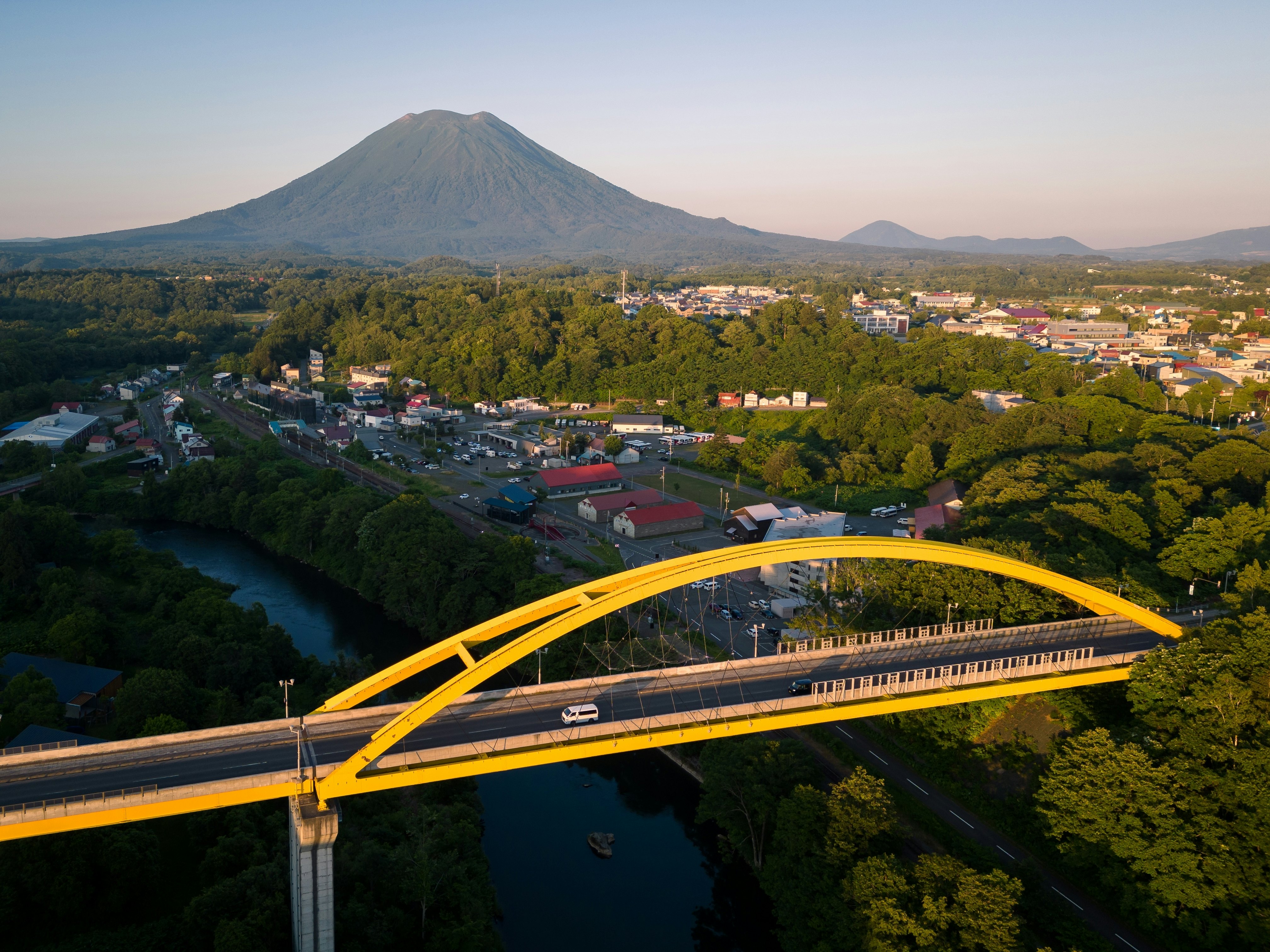 A van crossing a yellow bridge above a river with forest and a town in the distance and a volcanic mountain in the far distance on a sunny day.