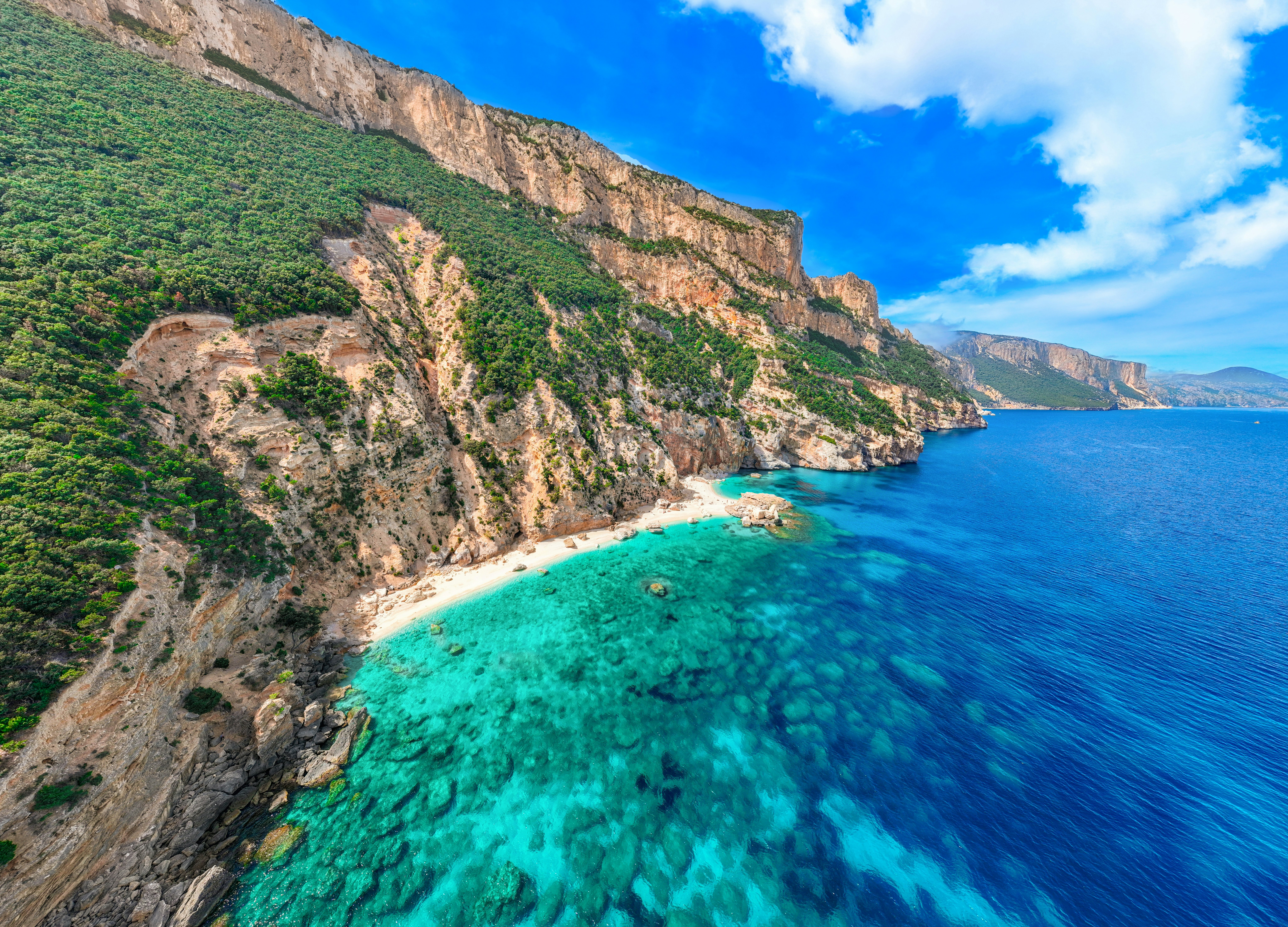 Aerial view of turquoise sea with rocky cliffs to the left and in the distance on a sunny day.