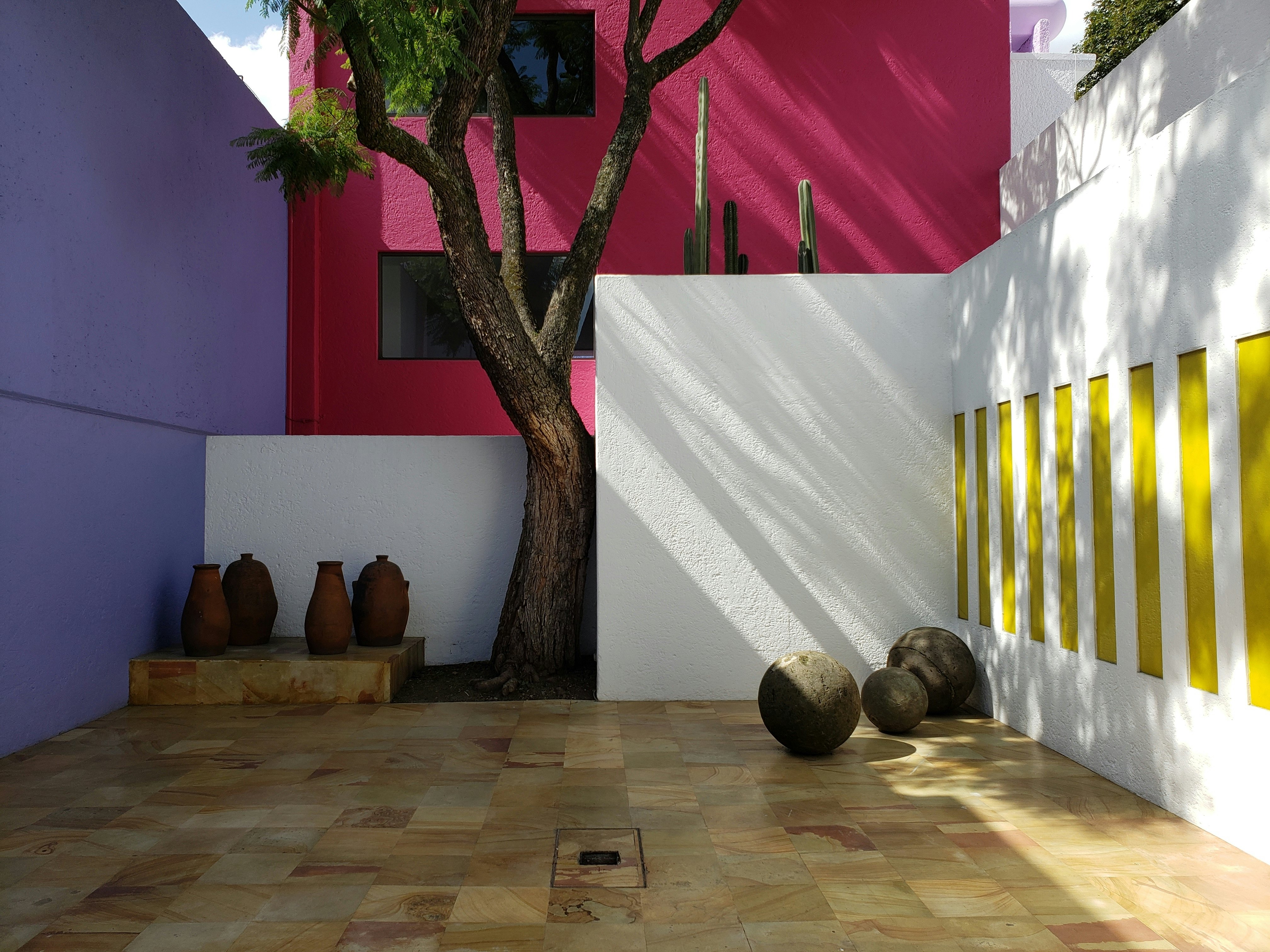 The courtyard of a home has one purple wall, one pink wall and a third wall with yellow panels over white. There are vessels on the ground and a small raised platform by a tree.