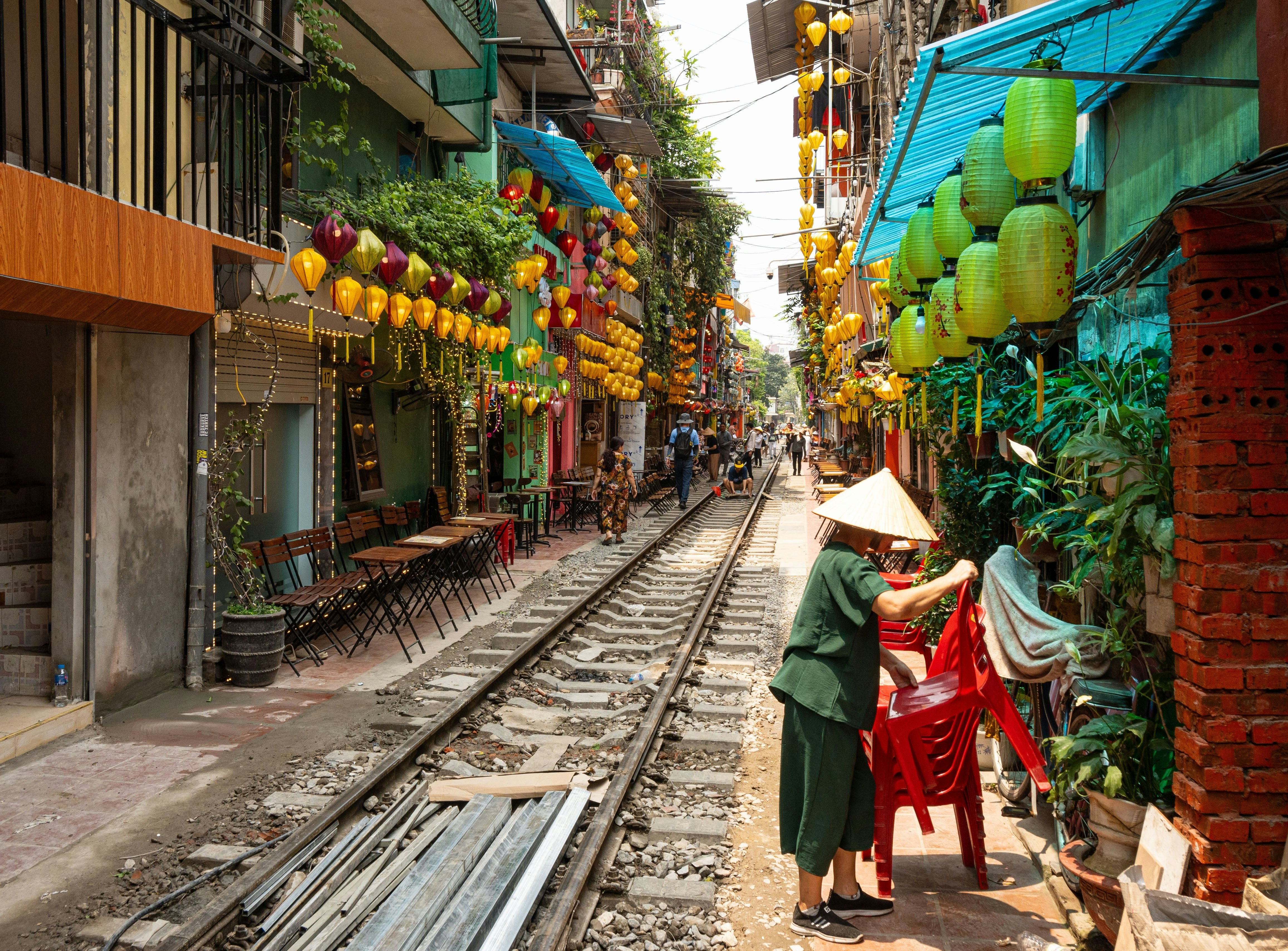 A woman stacks red plastic chairs on a street with train tracks and colorful balloons