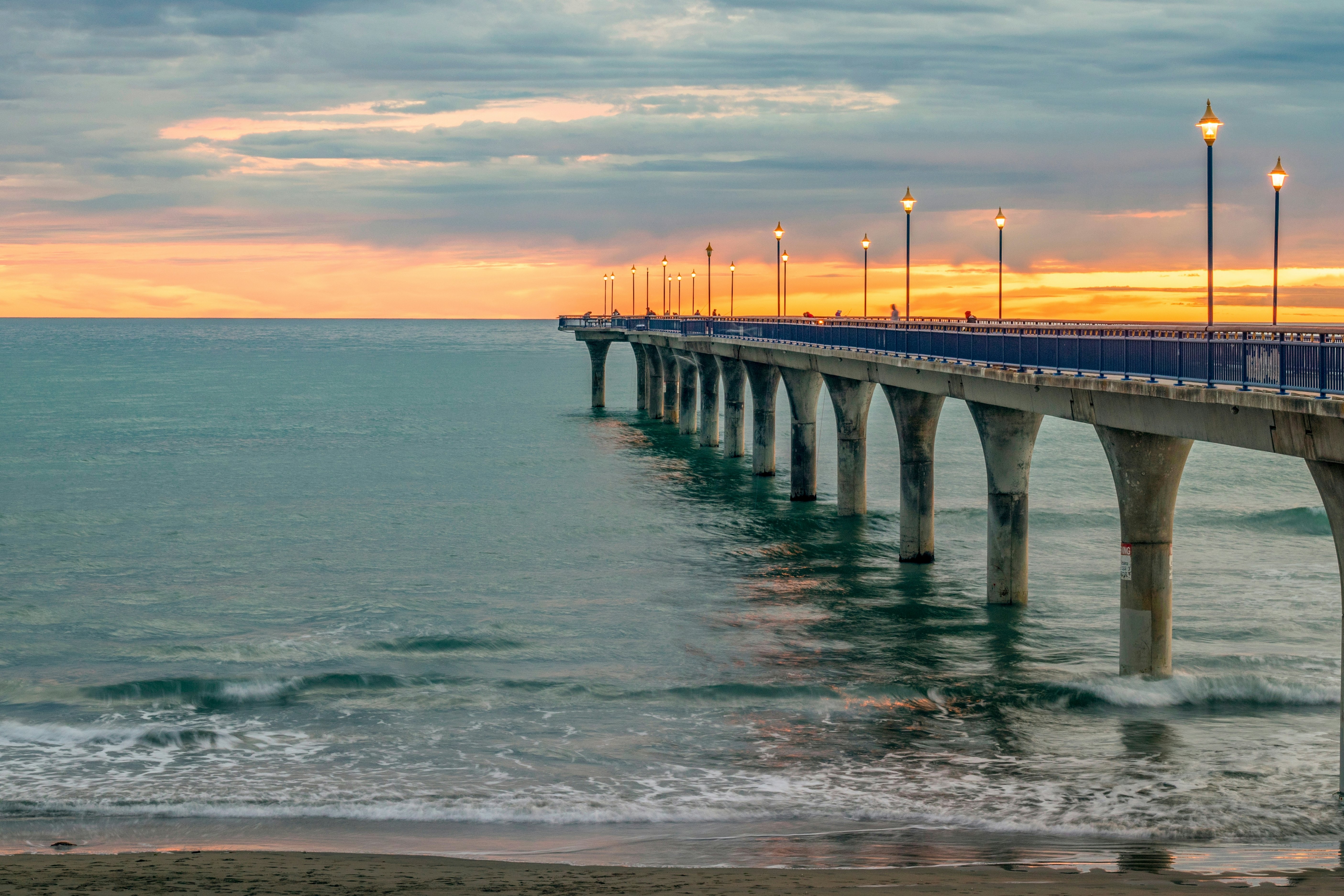 A long pier on concrete pilings with lighted poles along its walkway leading out into the sea at sunset.