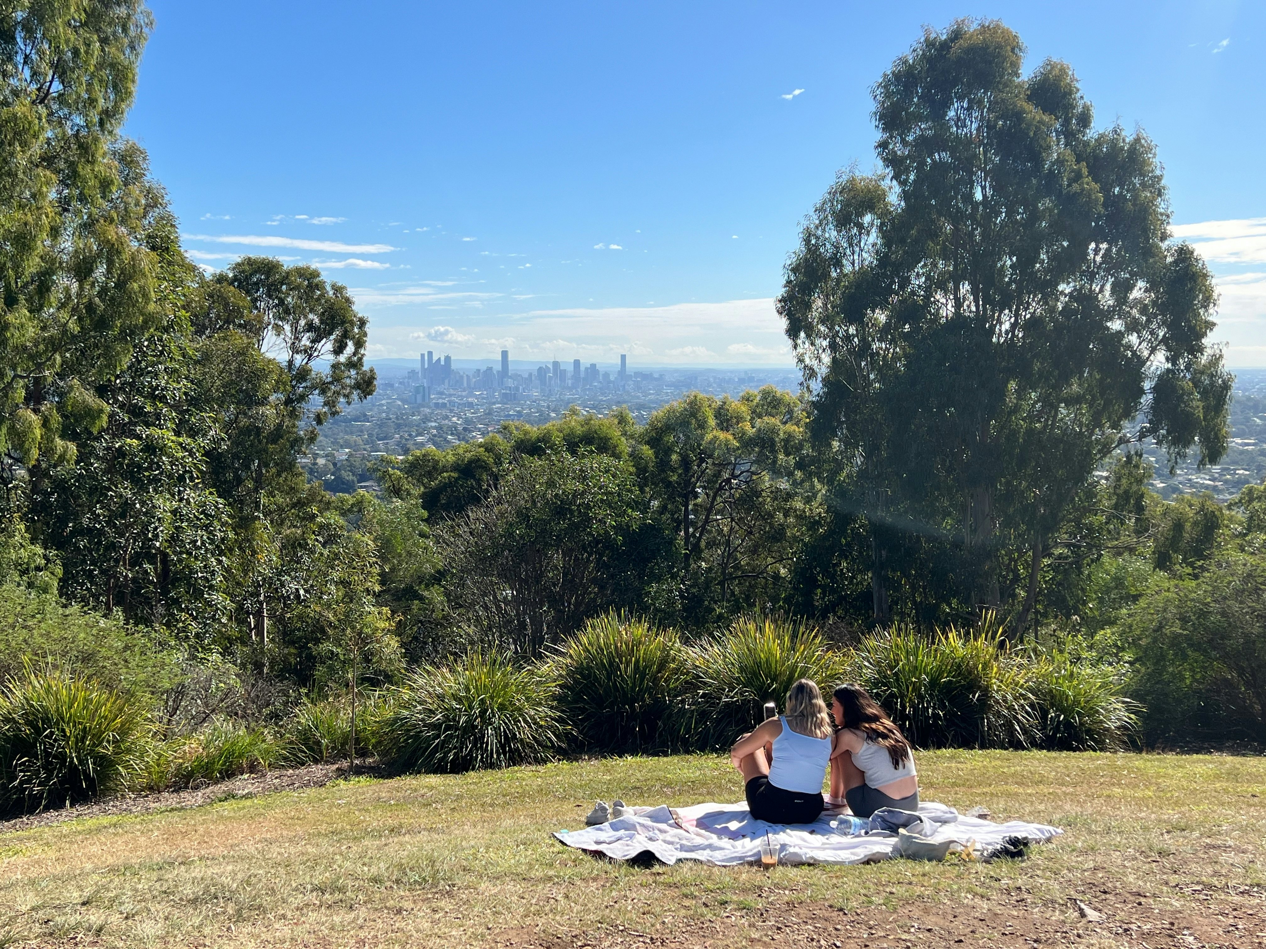 Two women sitting on a picnic blanket outdoors at a park viewing a city skyline in the distance on a sunny day.
