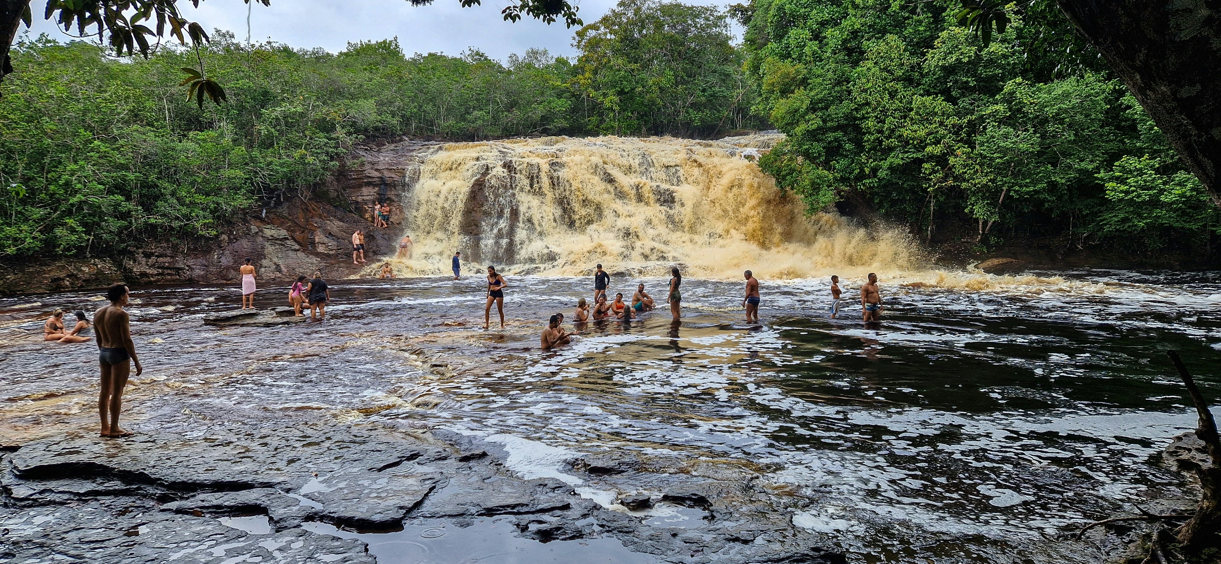 People in the water below a rushing waterfall surrounded by dense green vegetation.