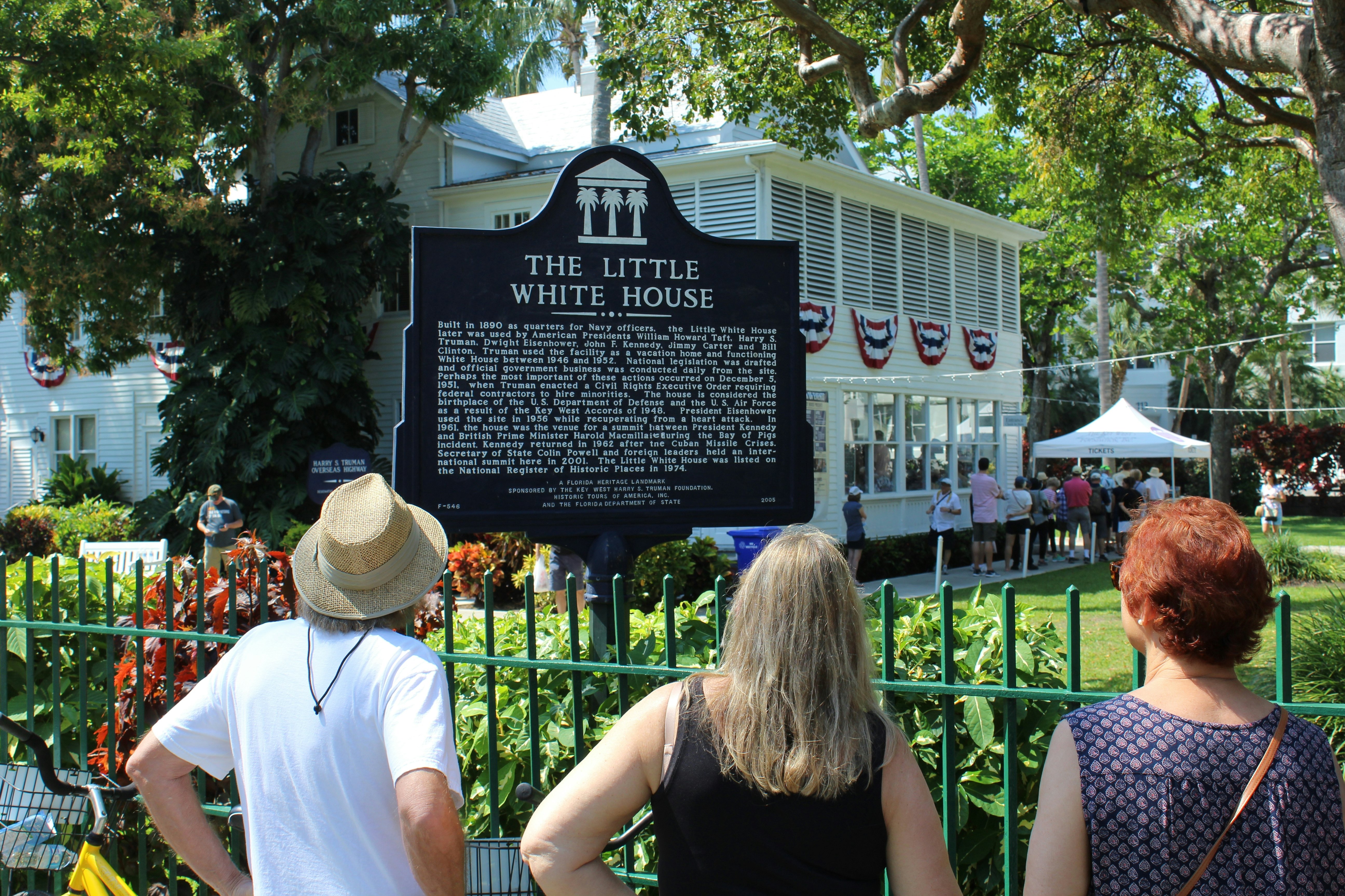 People read a historic marker by an iron fence in front of a white-painted house in a tropical location.