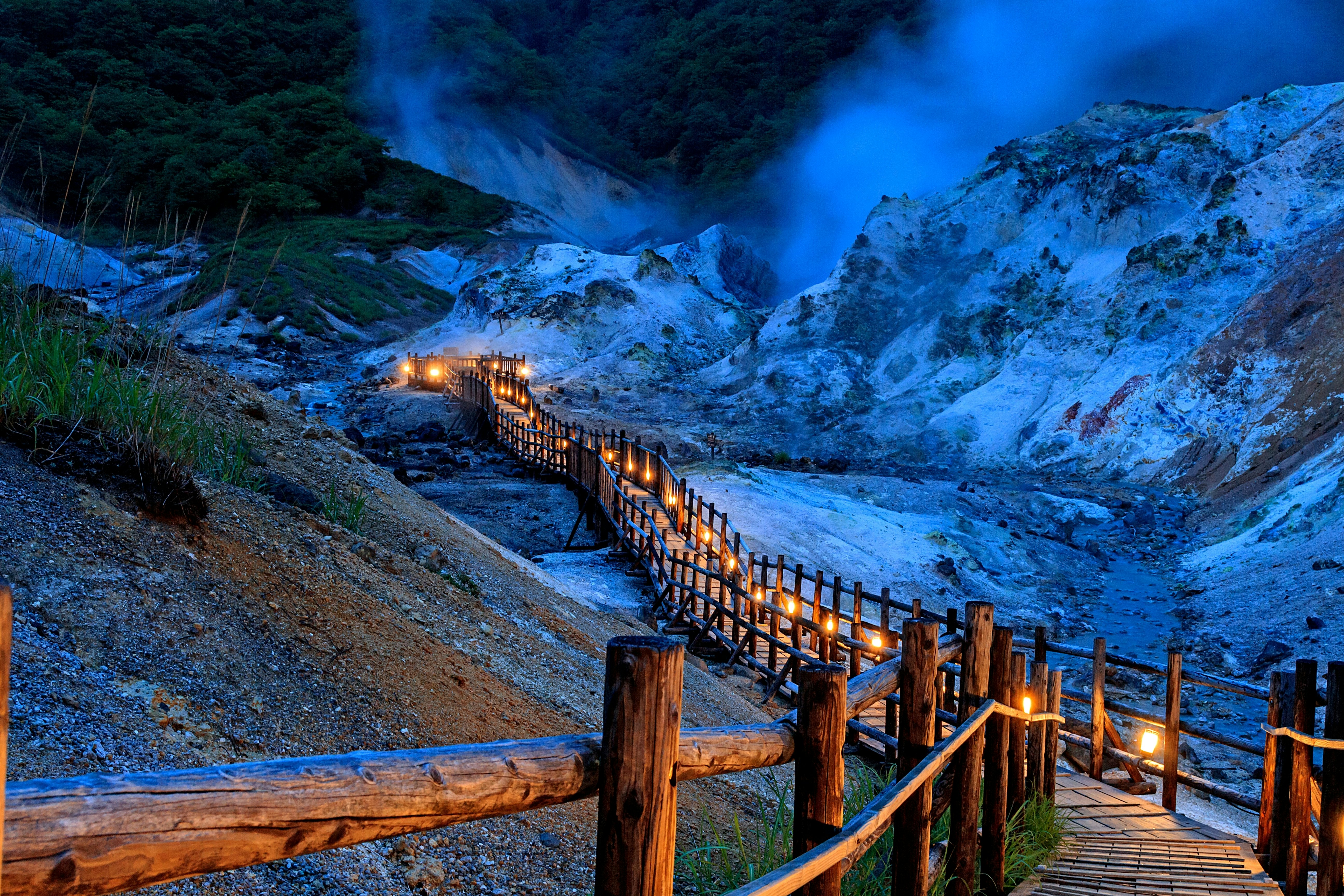An illuminated wooden boardwalk through a volcanic landscape at dusk.