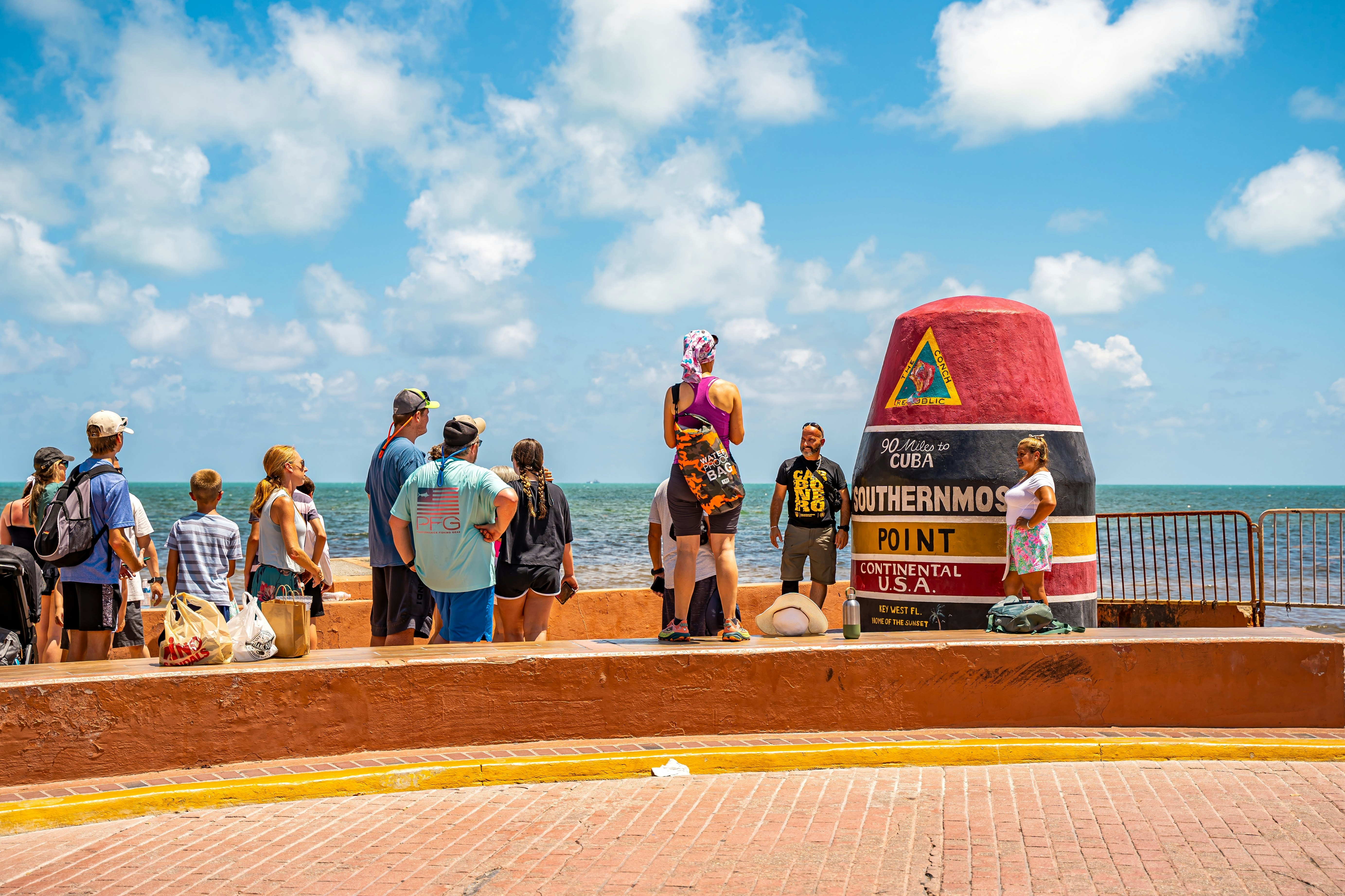 People line up to take photos in front of a large buoy-shaped marker by the sea.