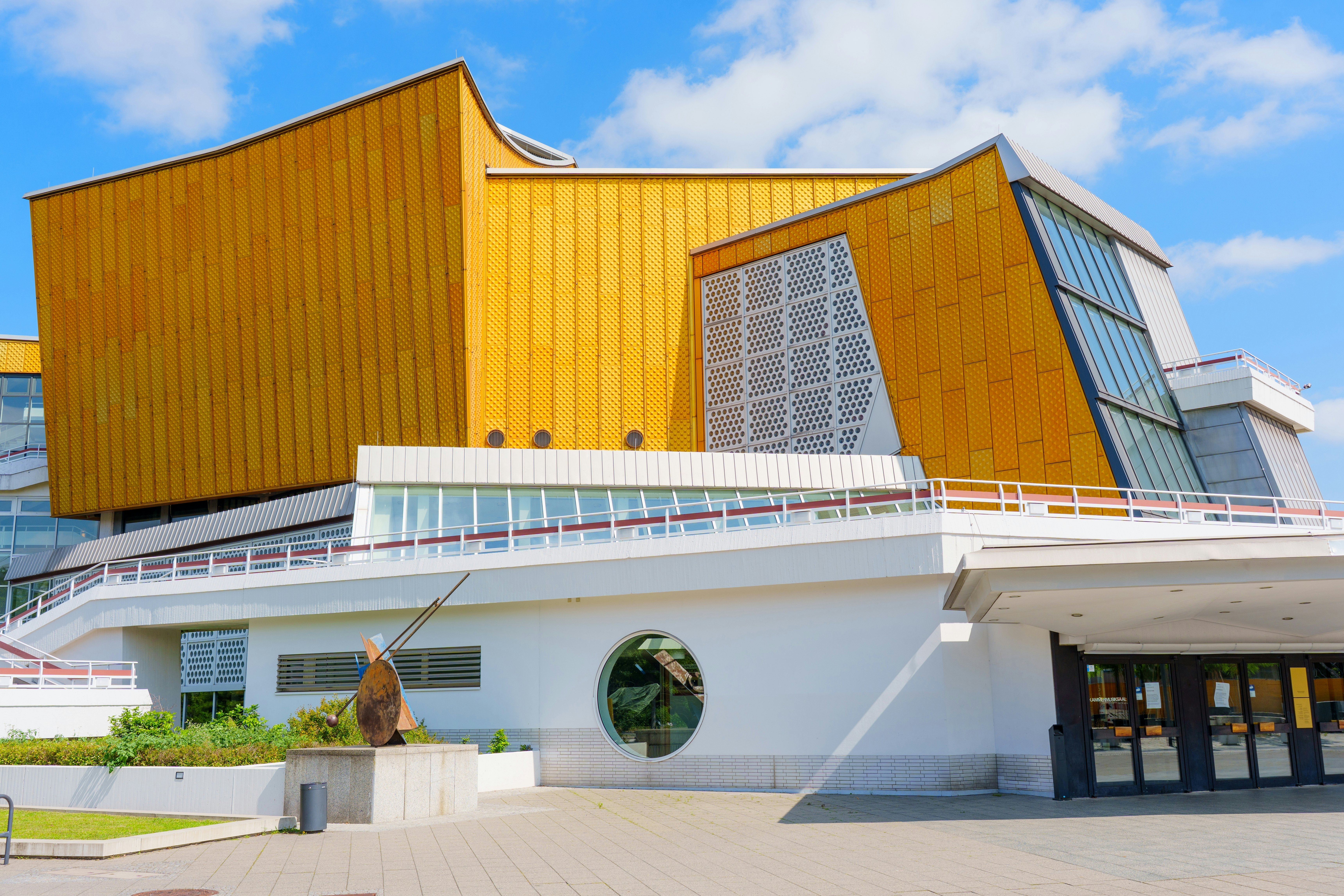 A large, modern building, white at the bottom and yellow at top, on a stone block plaza on a sunny day.
