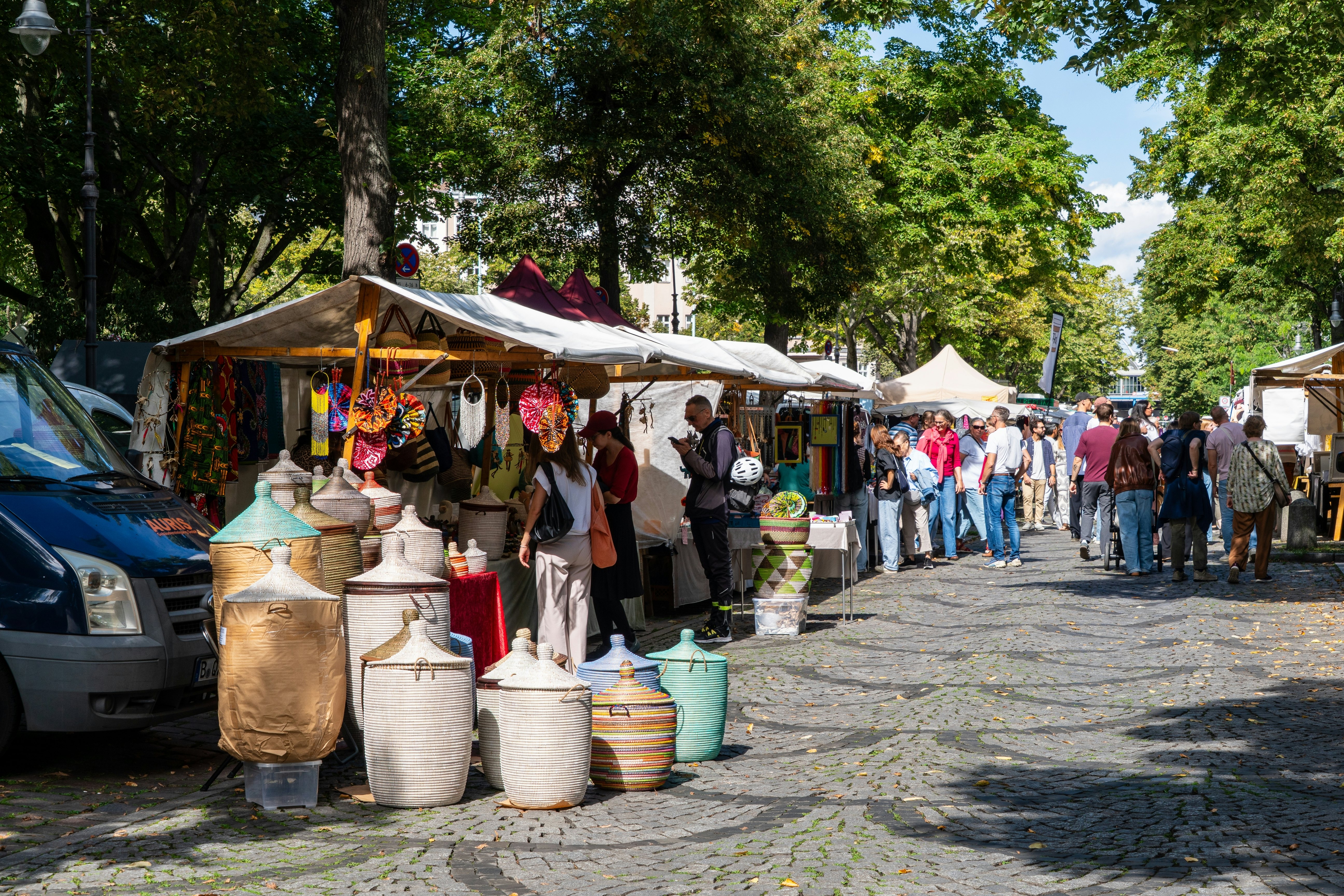 People shop at an outdoor market along a cobblestone street with tented vendor stalls on a sunny day.