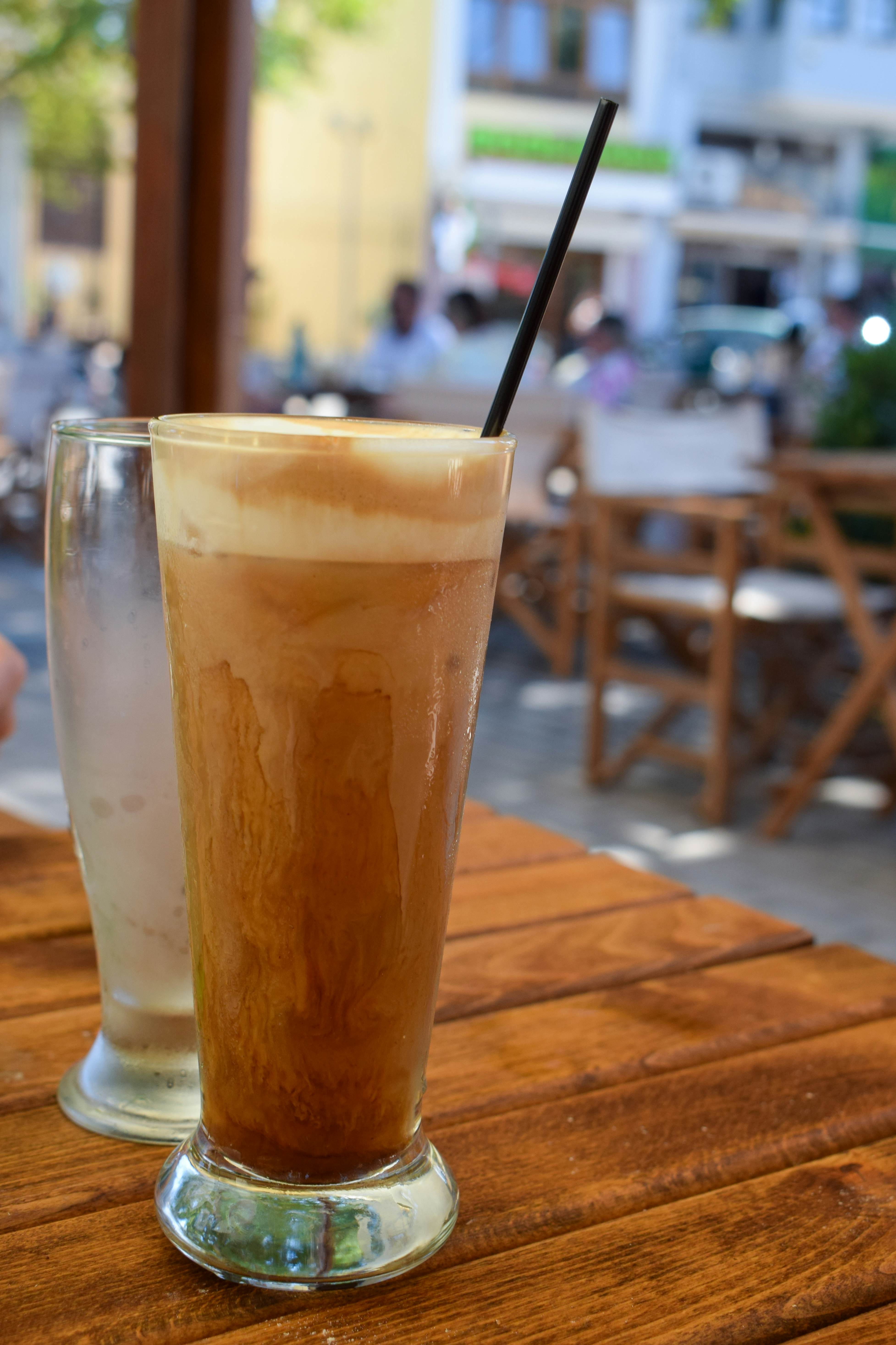 A tall clear glass with a coffee drink with foam on top on a wooden table.