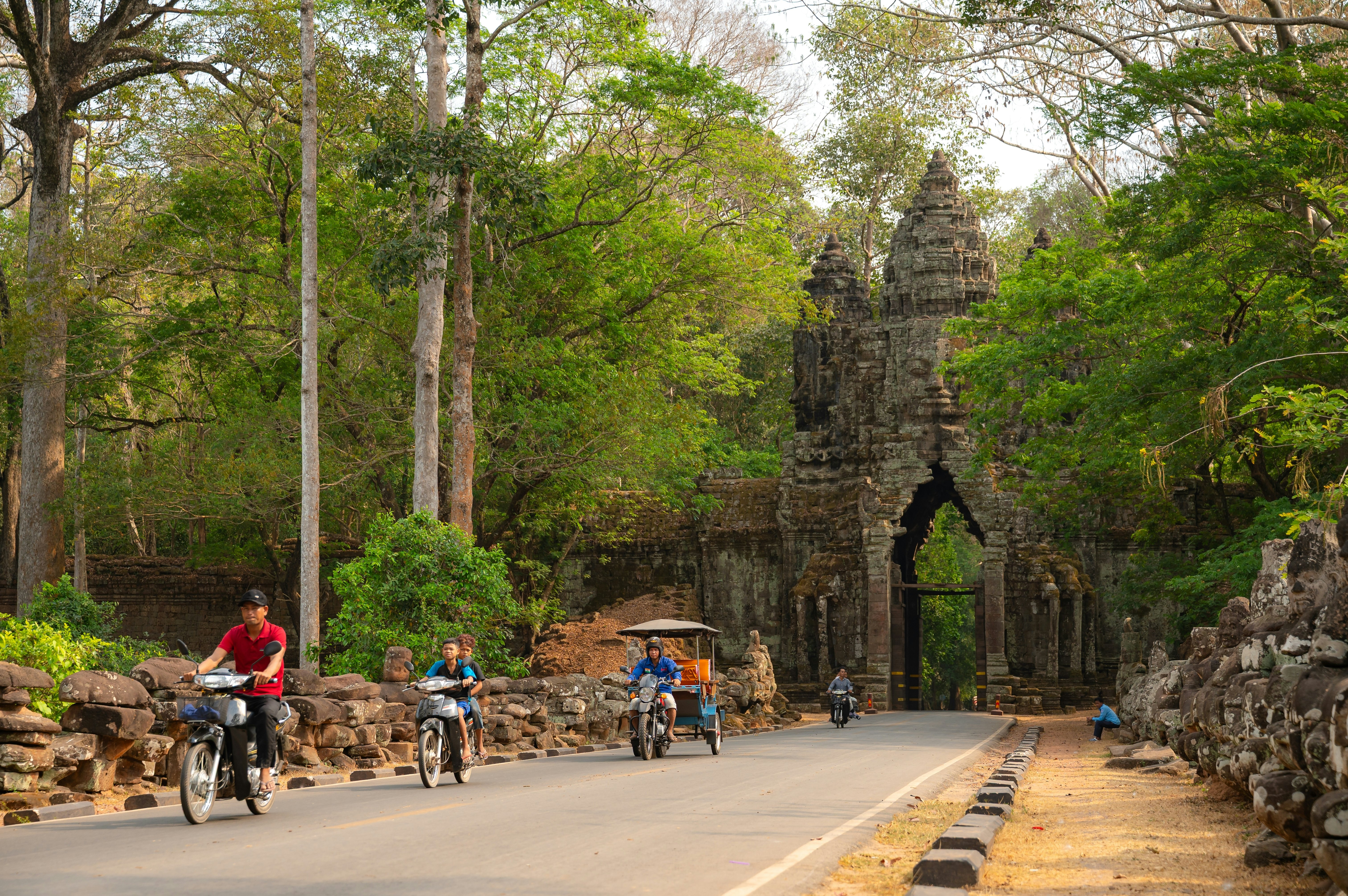 Bicyclists and rickshaw drivers on a paved road that leads to the stone gate of a temple complex in a forest.