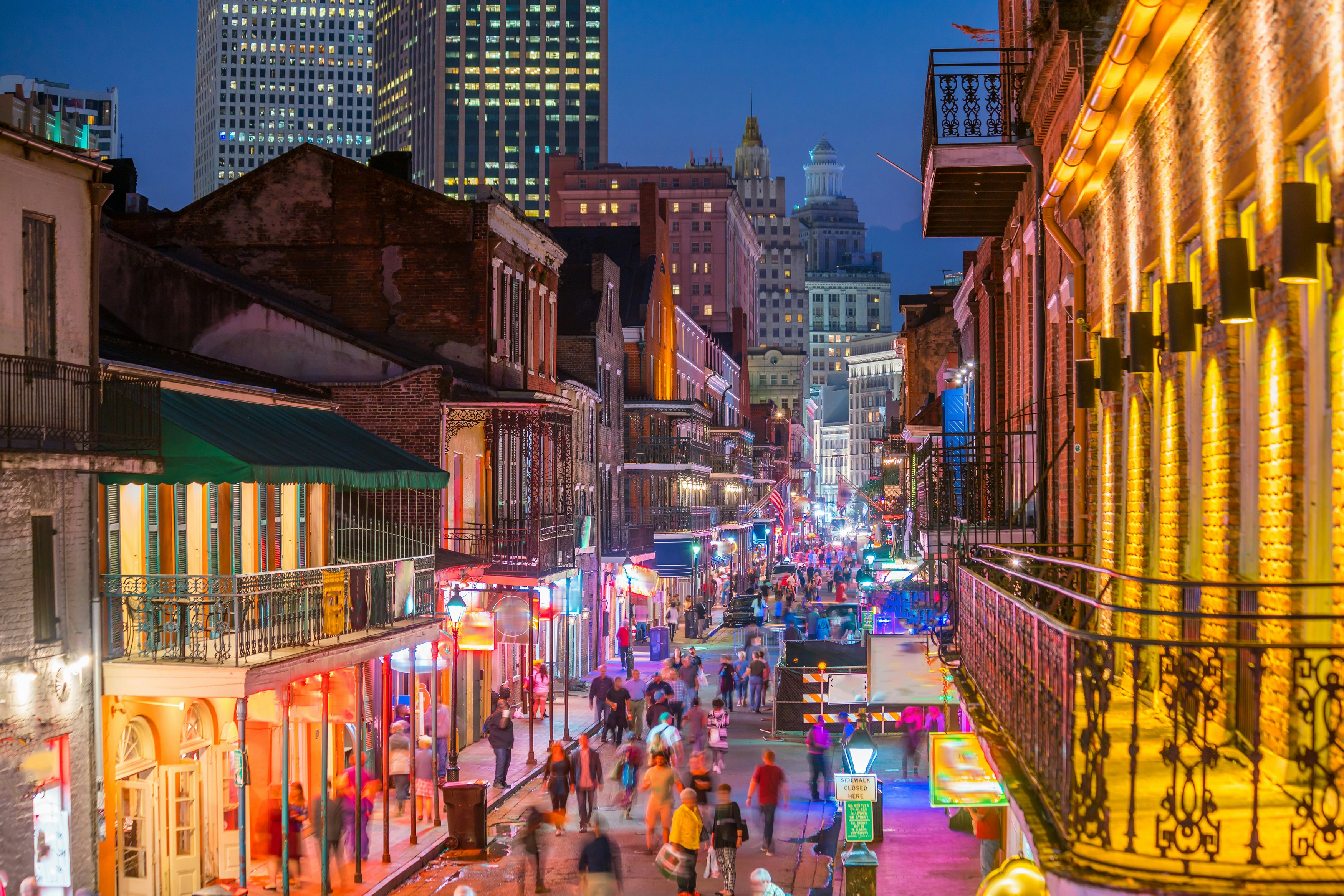 A pedestrianized street is lit by colored lights and has numerous bars. Skyscrapers are in the distance behind the historic buildings of the neighborhood.