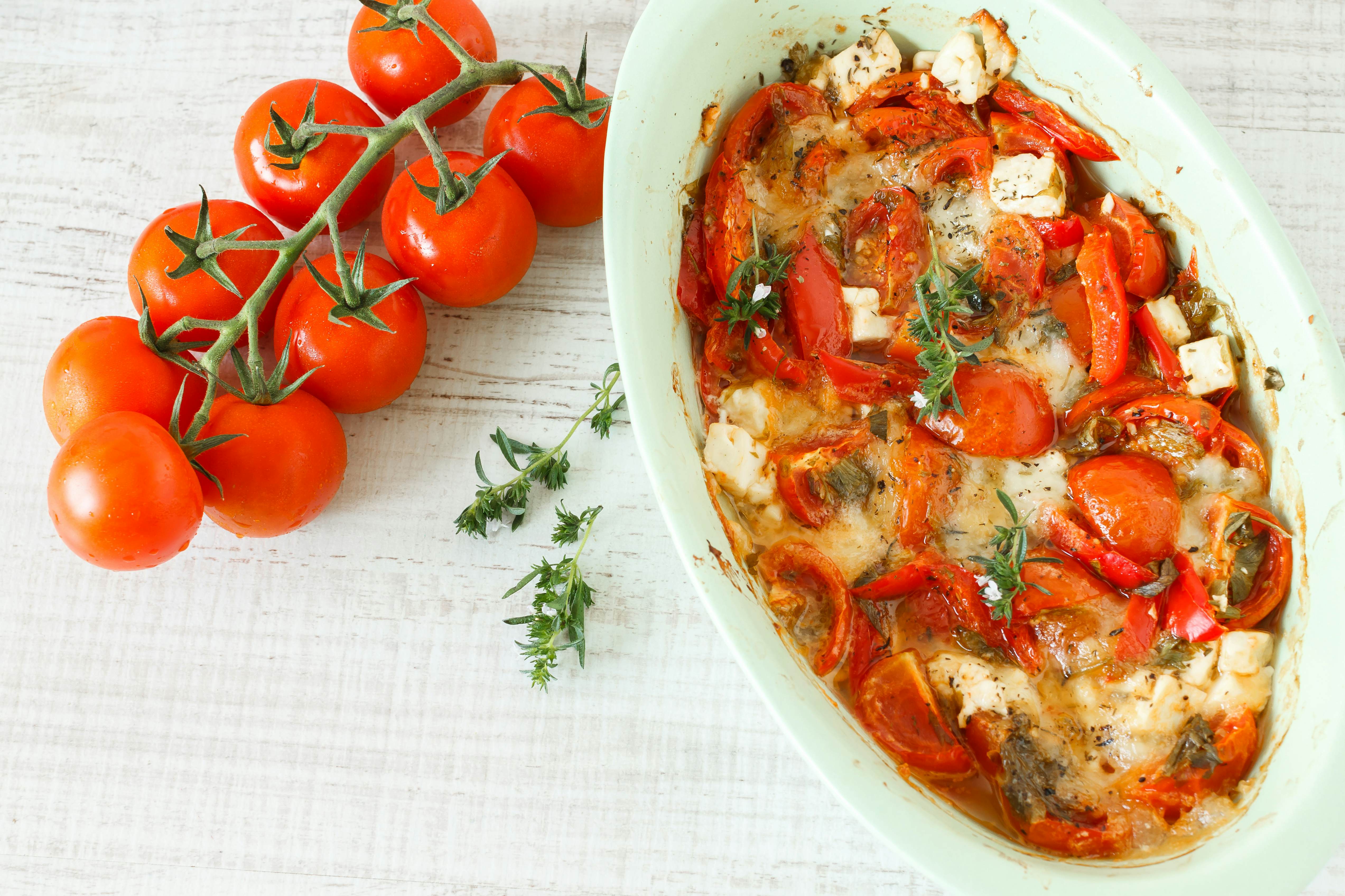 A vine with fresh red tomatoes and a casserole dish with squares of cheese topped with red peppers and green herbs.