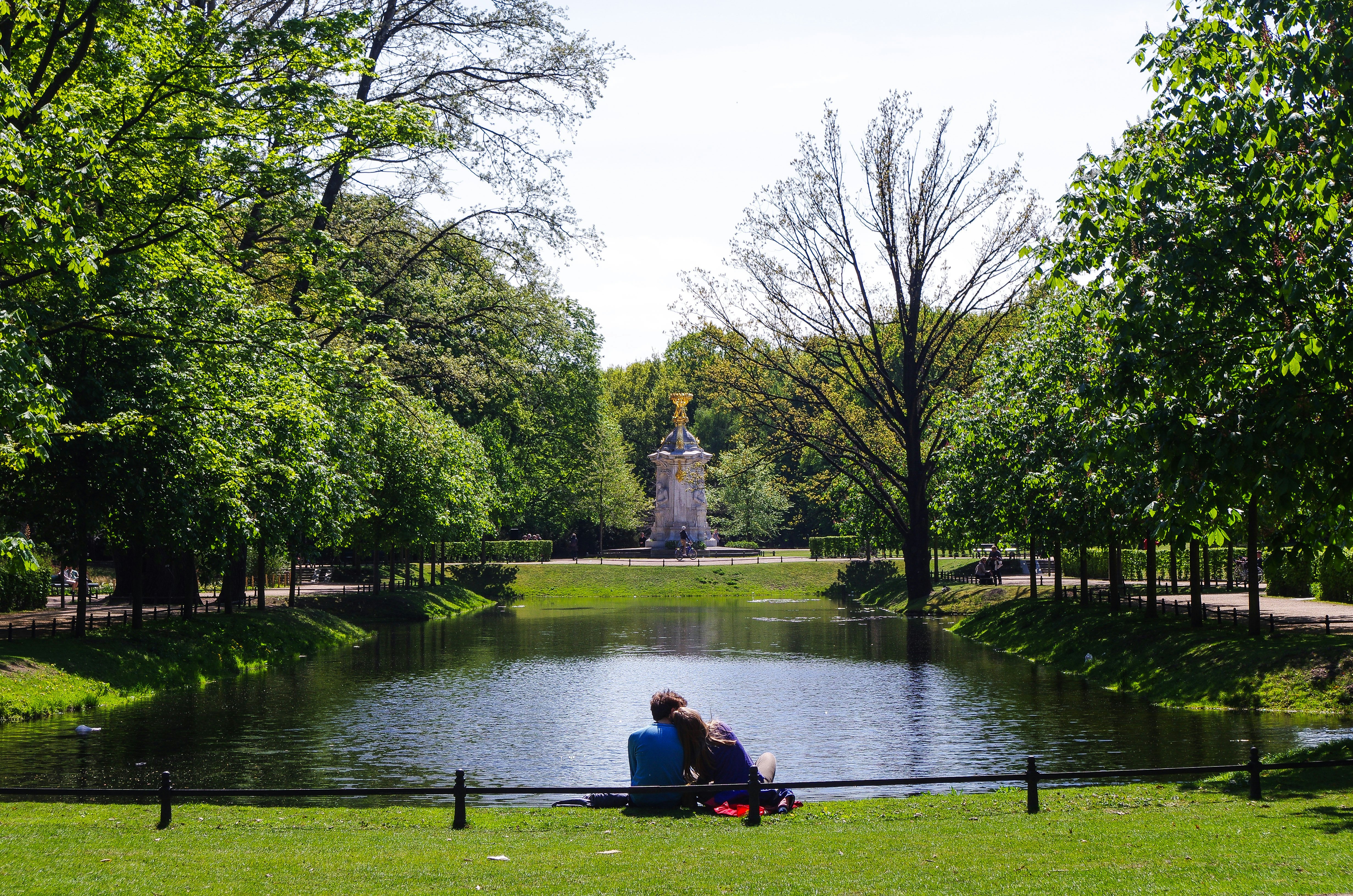 A couple sat together by a pond in green parkland.
