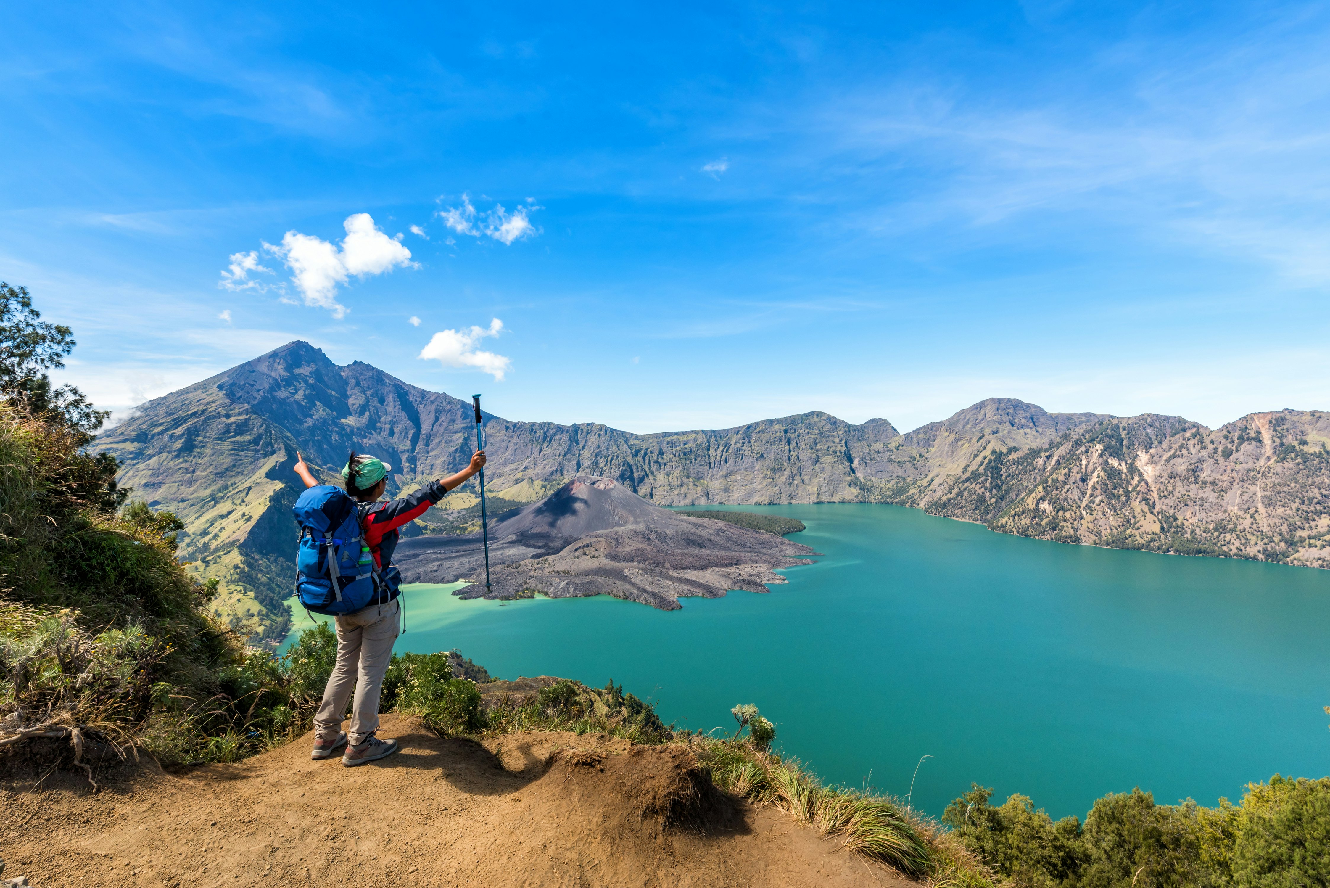 A hiker with arms outstretched holding a hiking pole looks down at a blue lake with rocky mountains on the far shore.