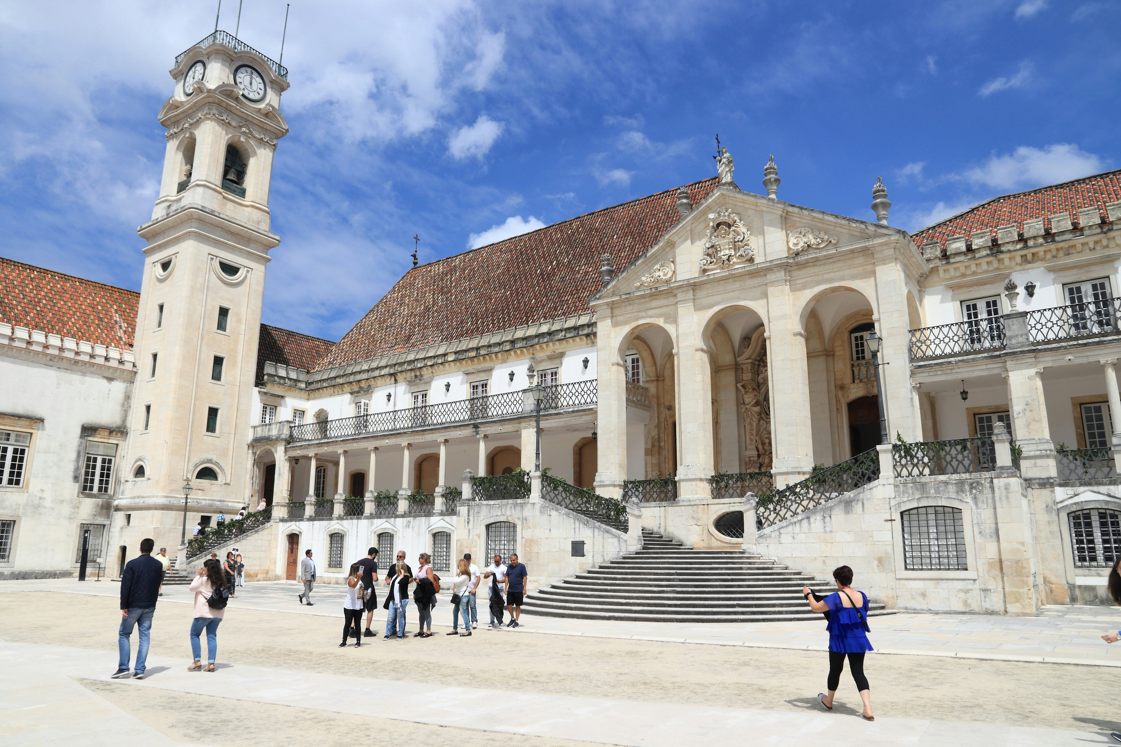 People touring white flagstone courtyard near stone buildings with reddish roofs on a sunny day.