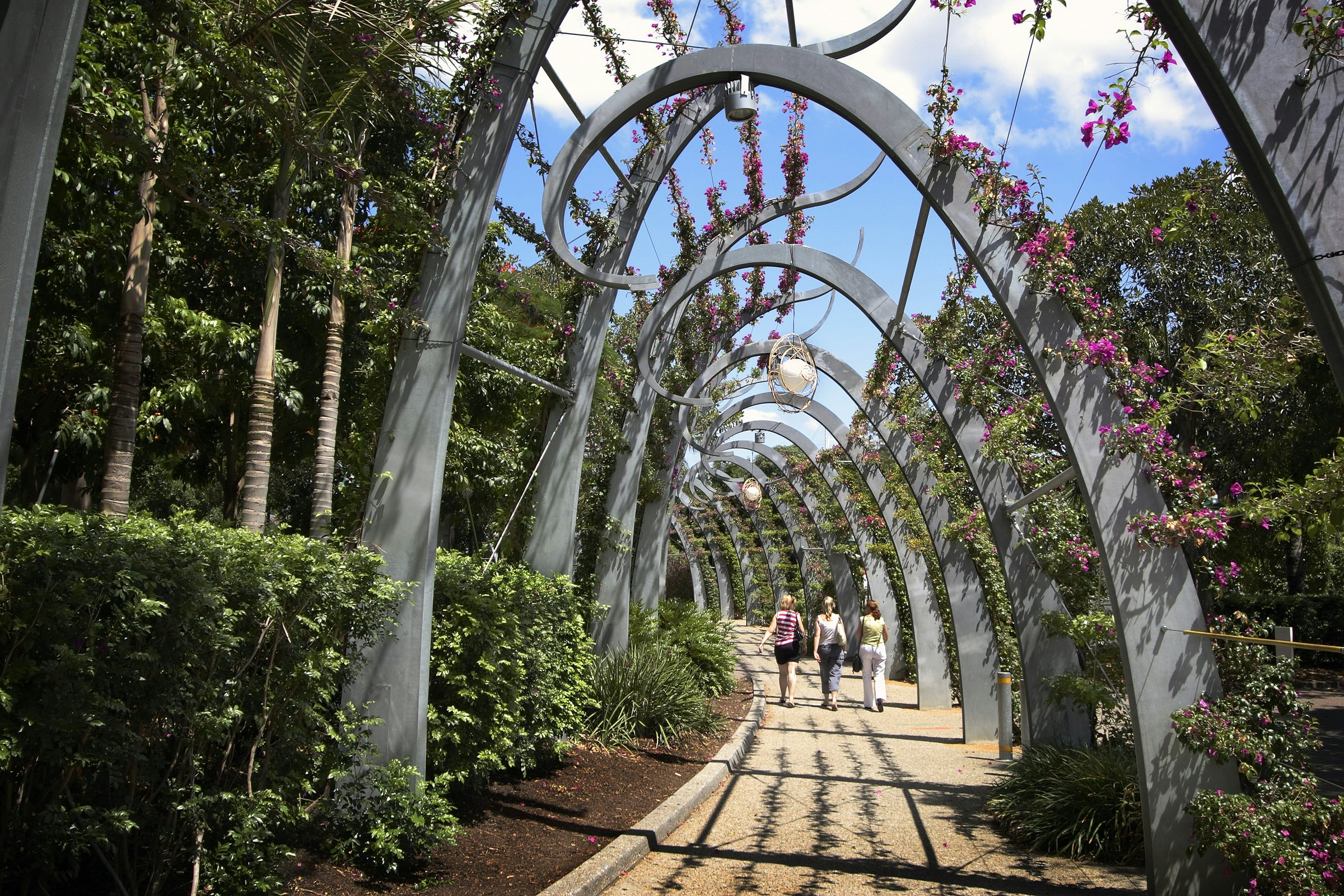 Three women walk along a stone path covered in modern, arched trellis with flowers above on a sunny day.