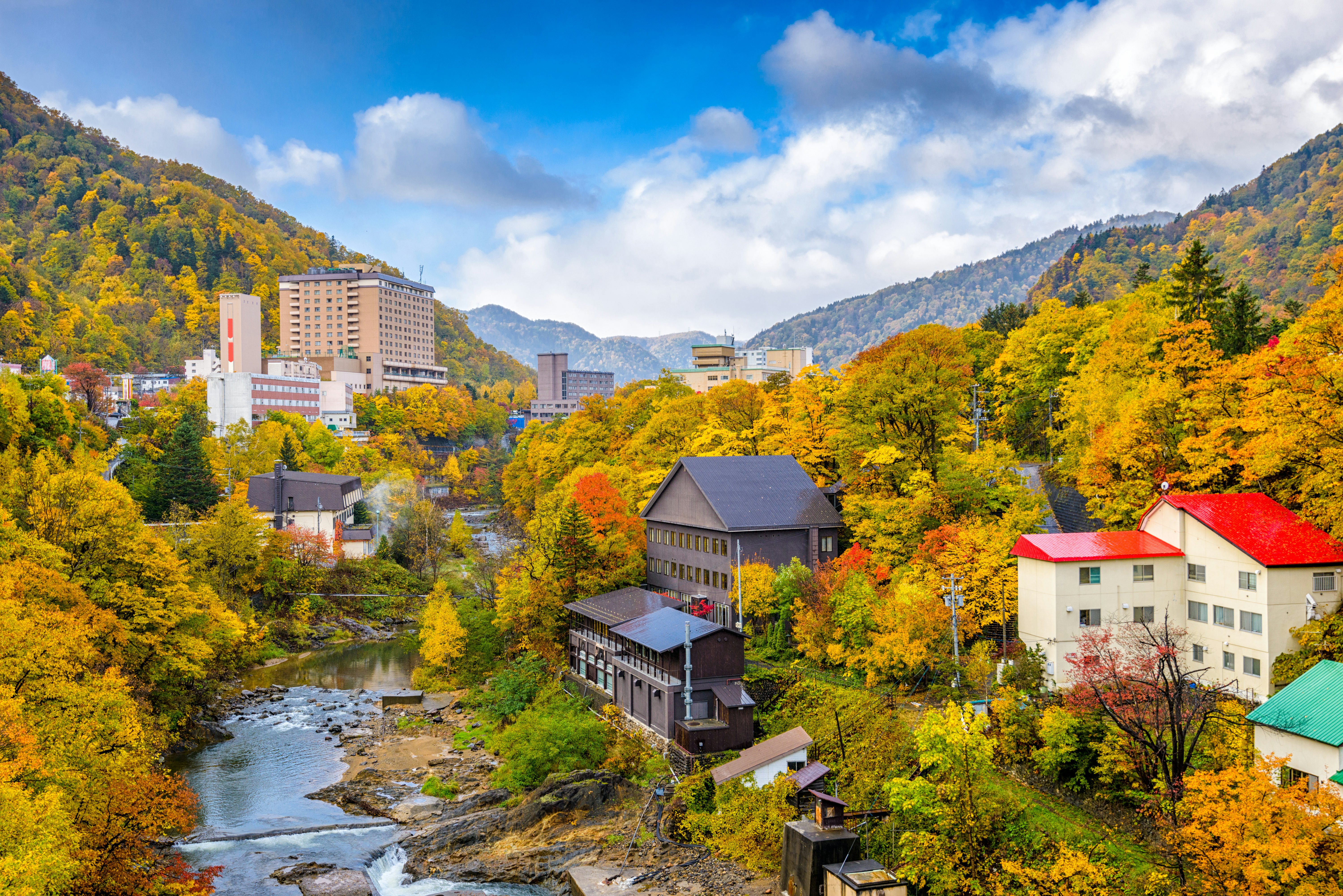 Buildings along a narrow river surrounded by trees with bright orange and yellow leaves on a sunny day.